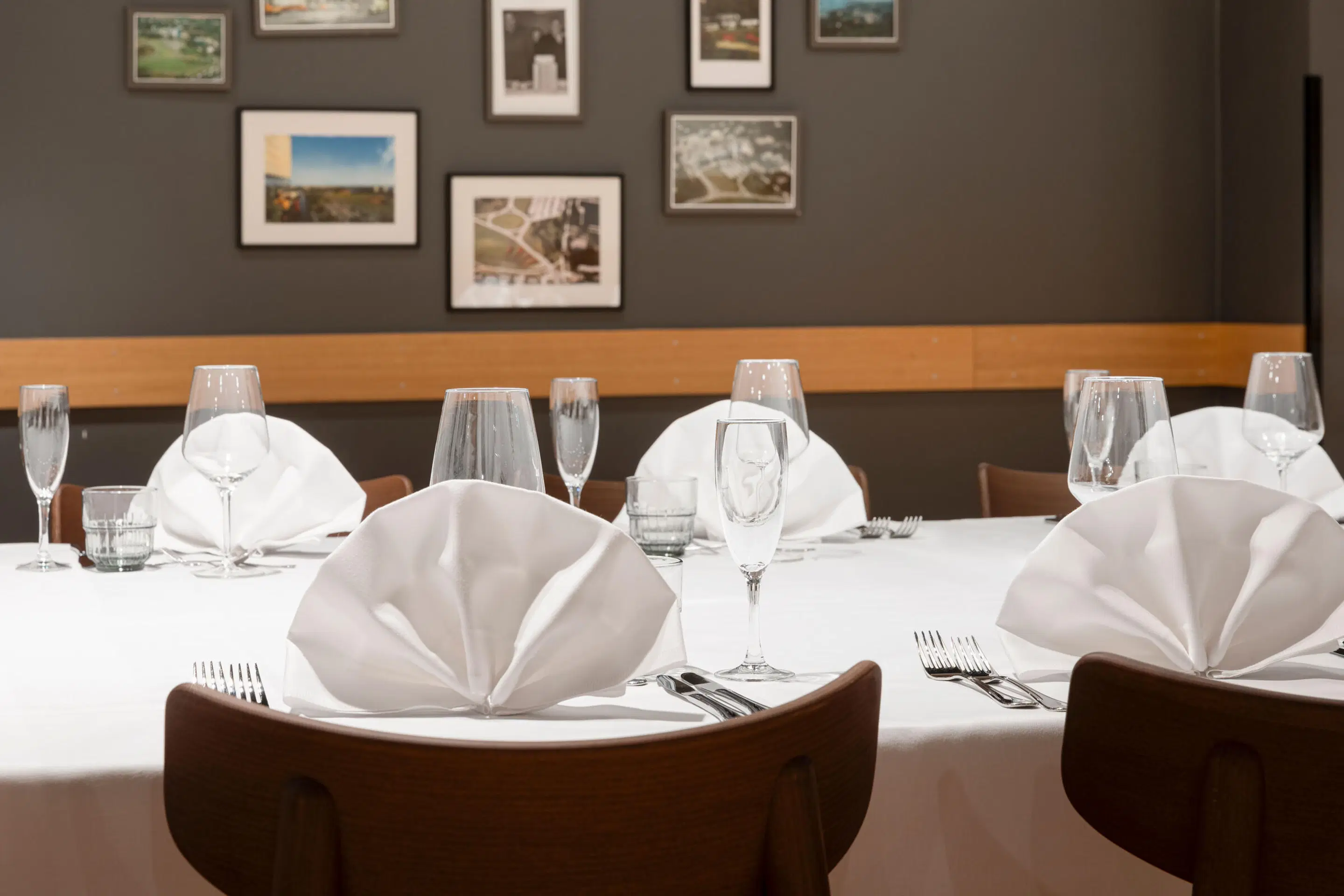 Elegantly set dining table with folded white napkins, wine glasses, and cutlery. Dark wall in background displays framed photographs.