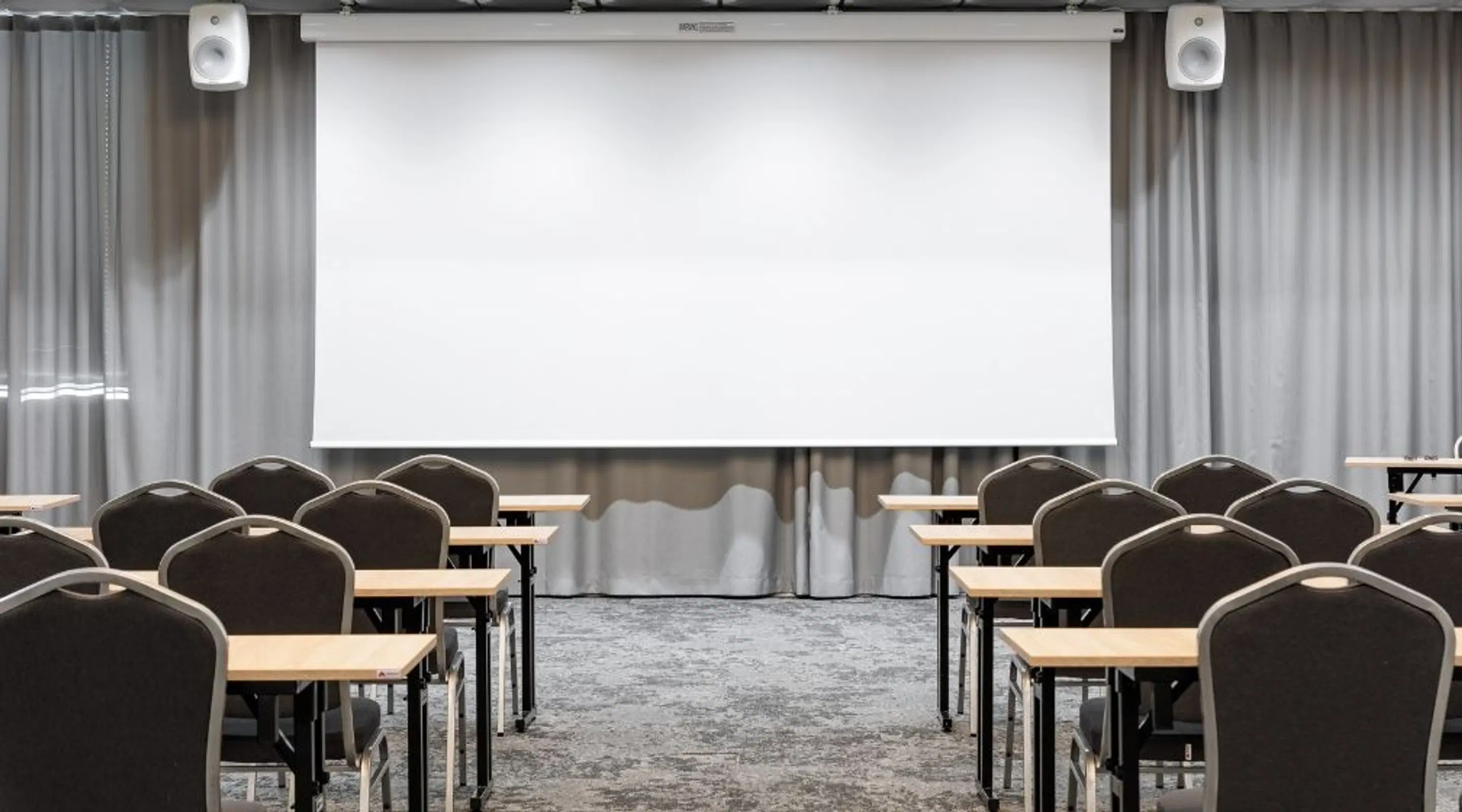 Konverentsiruum, kus lauad ja toolid on asetatud ridadesse projektsiooniekraani ees. Hall vaipkate ja helehallid kardinad.