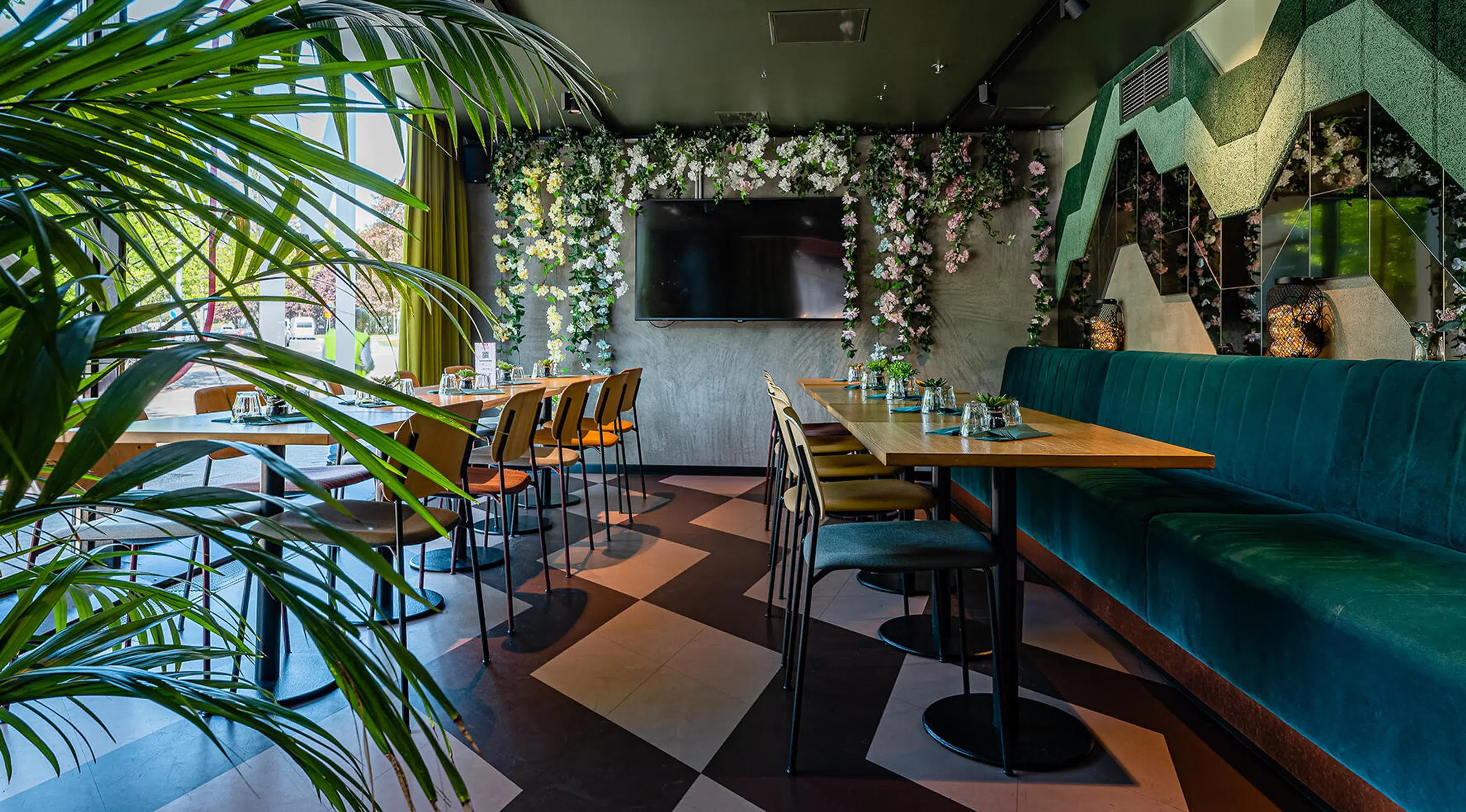 Restaurant dining area with wooden tables and chairs. Space decorated with plants and floral adorned walls. Hard tile flooring with geometric pattern.