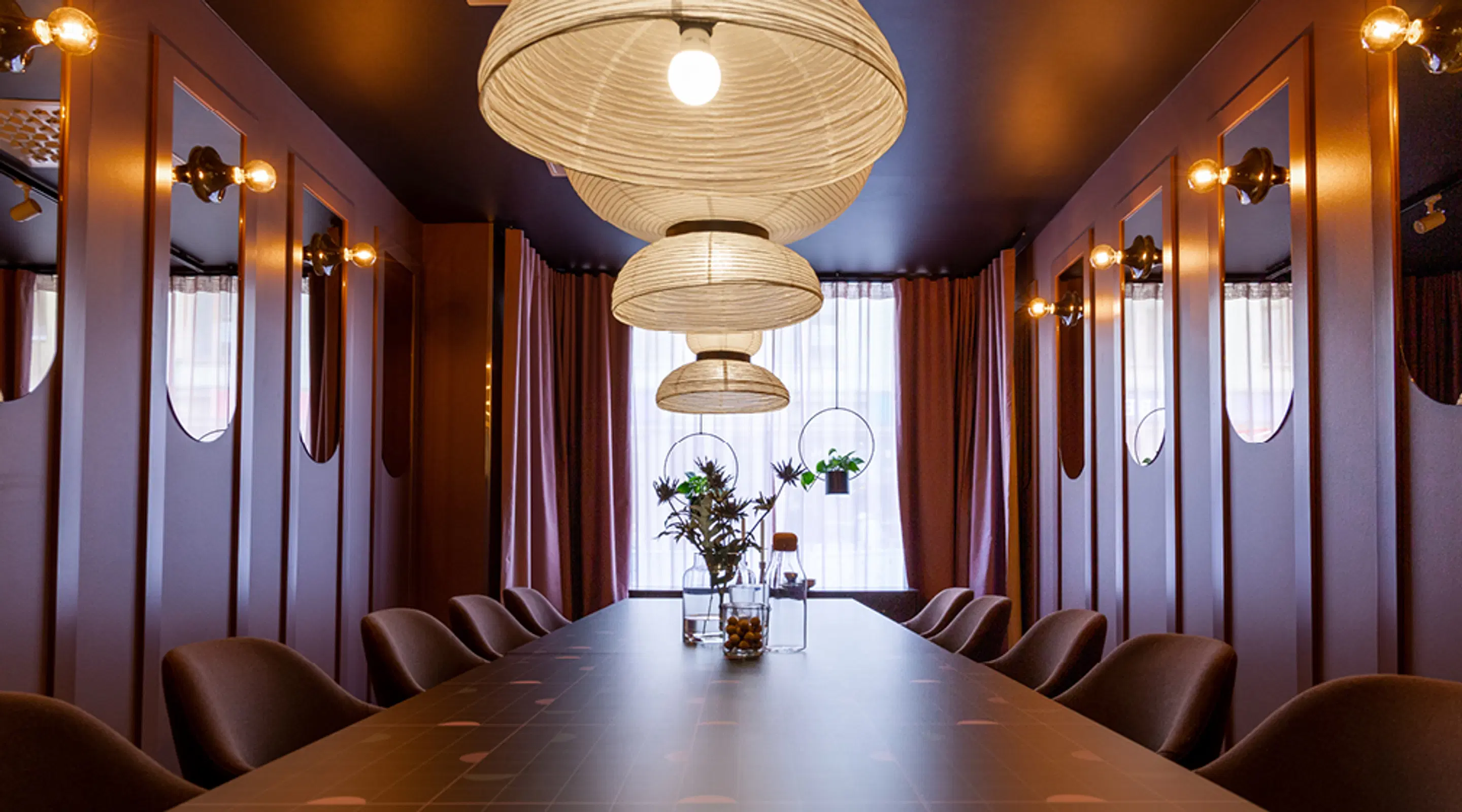 Stylish meeting room with long wooden table, brown chairs, hanging pendant lights and potted plants by the window.