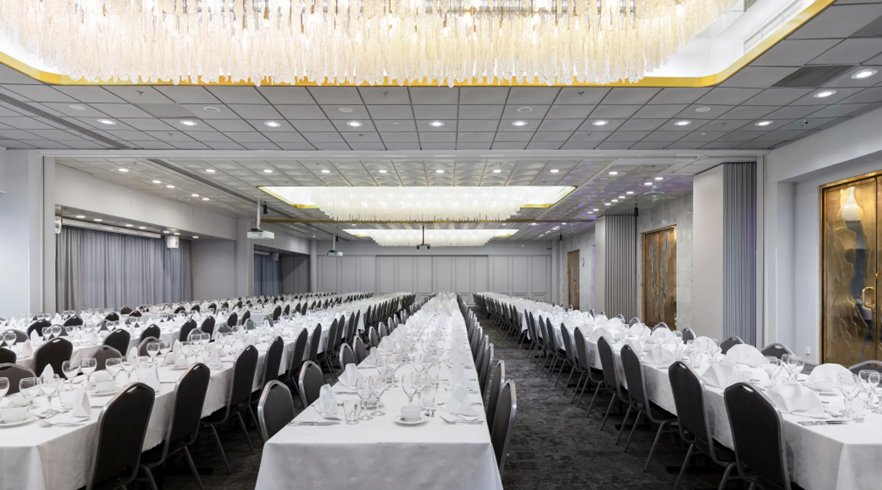 Spacious banquet hall with long tables covered in white tablecloths and black chairs. Crystal-like chandeliers hang from the ceiling.