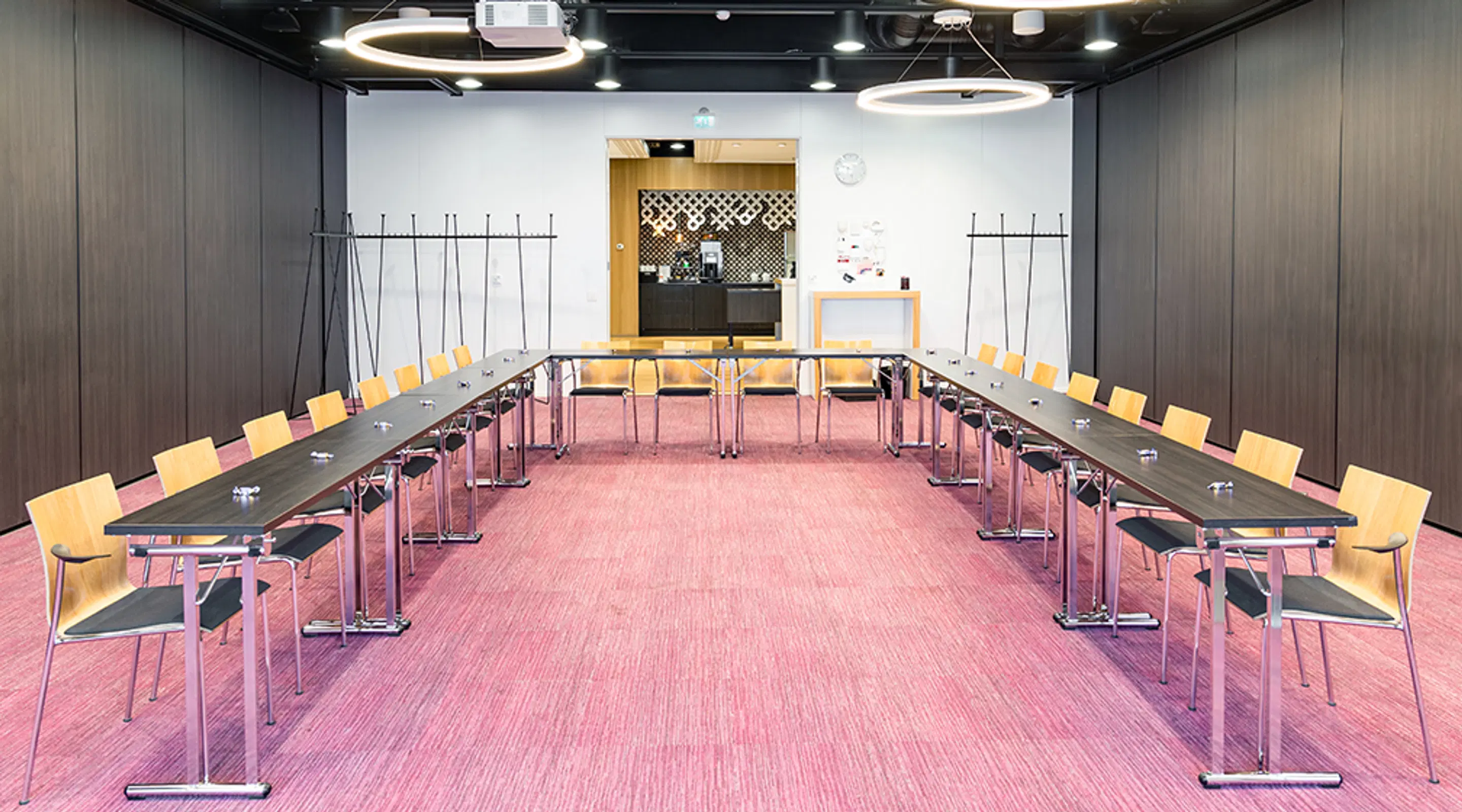Modern meeting room with U-shaped table arrangement, wooden chairs and pink carpeted floor. Wide pathways and level floor surface.