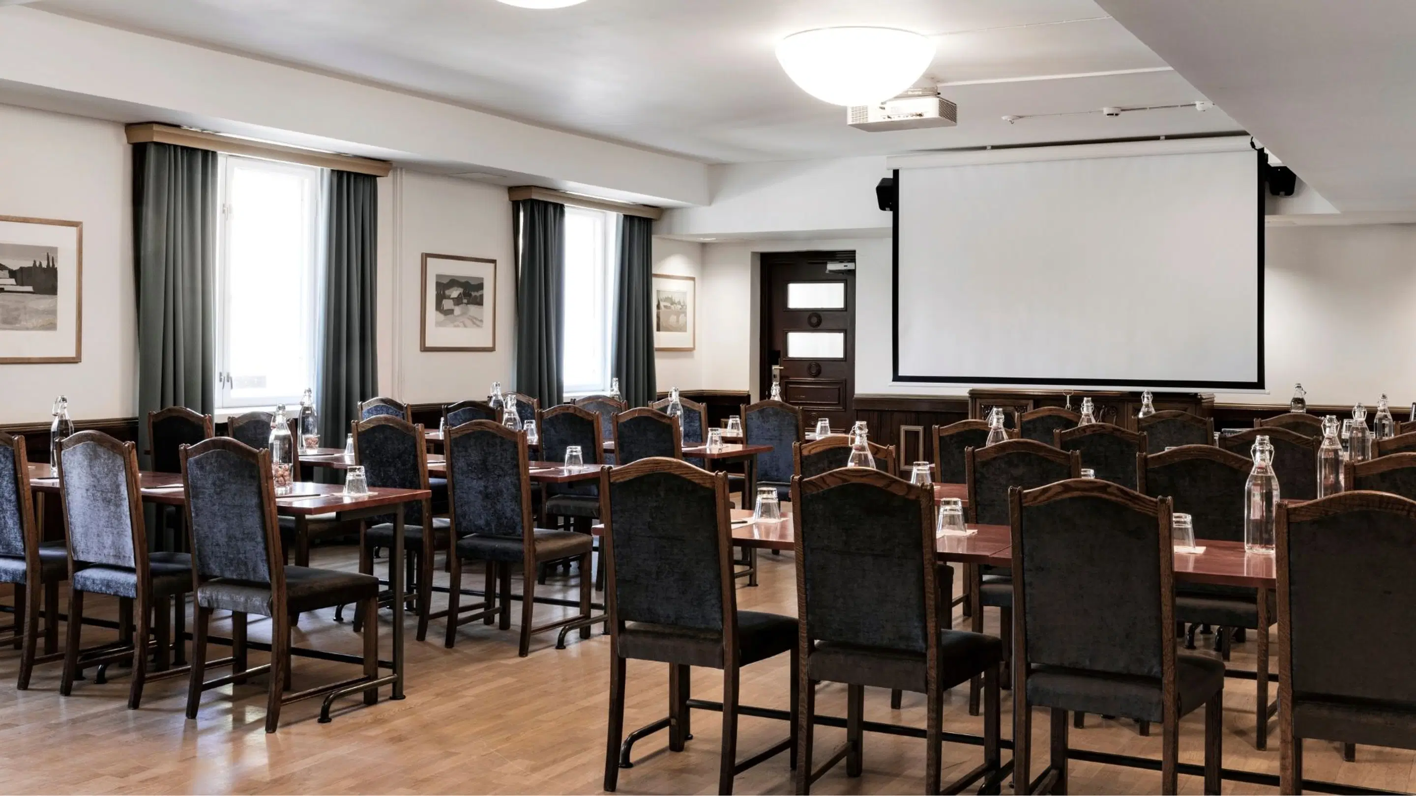 Conference room with tables and chairs in classroom layout, projection screen at front, wooden flooring and large windows with dark curtains.