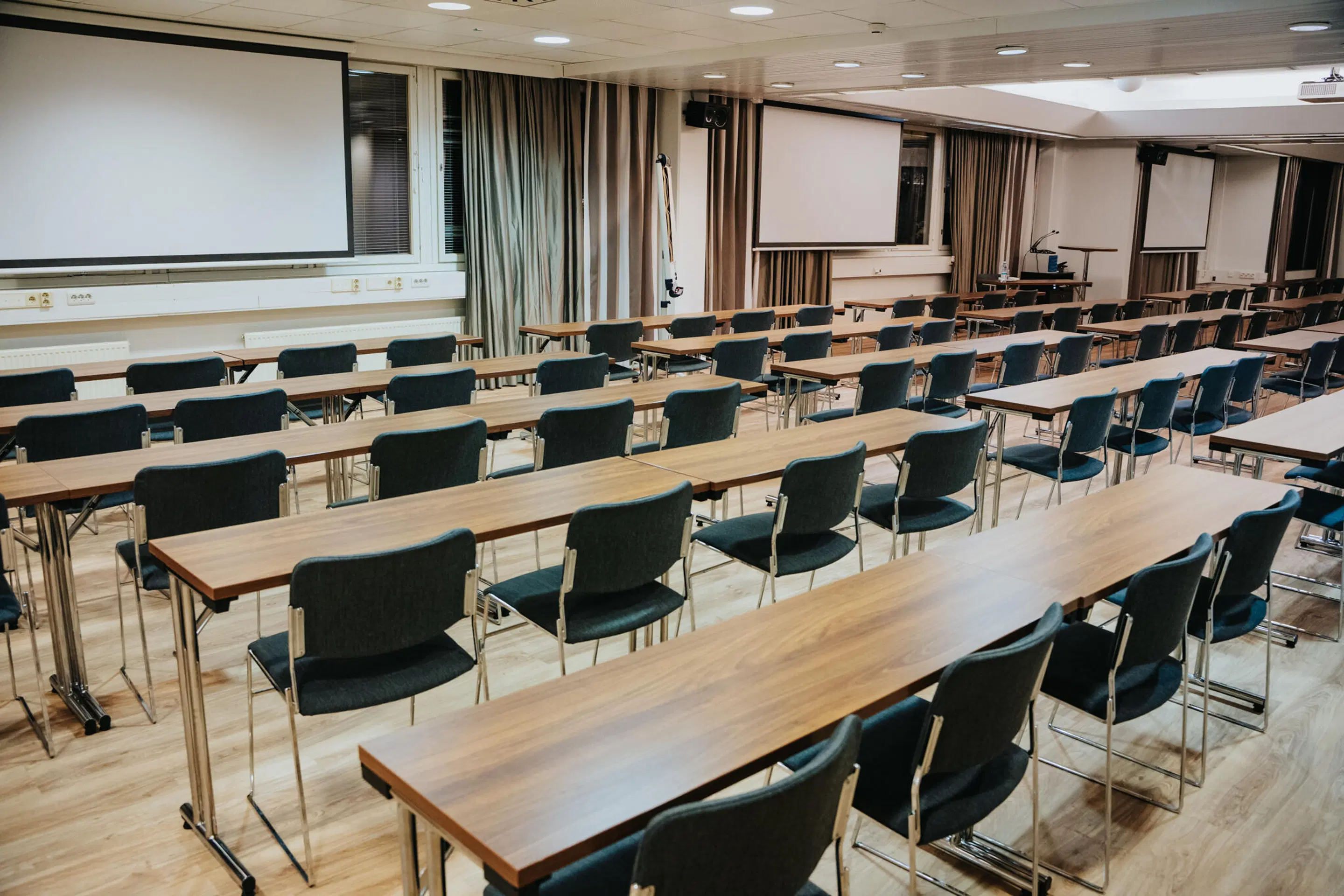 Auditoorium pikkade laudade ja toolidega ridades. Ees ekraanid ja taustal kardinad. Tasane puitpõrand ja hea valgustus.