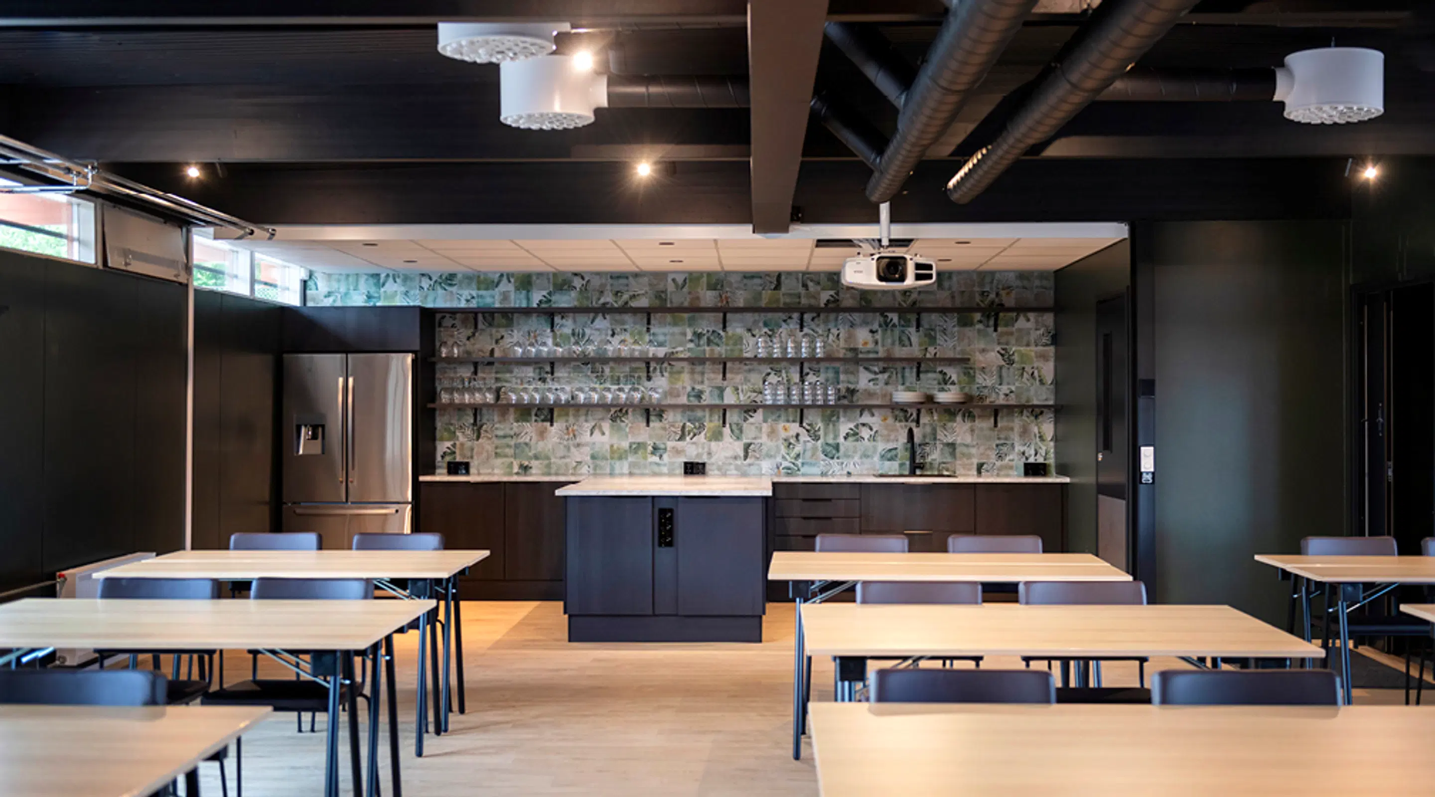 Modern meeting room with wooden tables, dark kitchenette in background. Ceiling-mounted projector, light wooden floor and industrial ceiling structures.