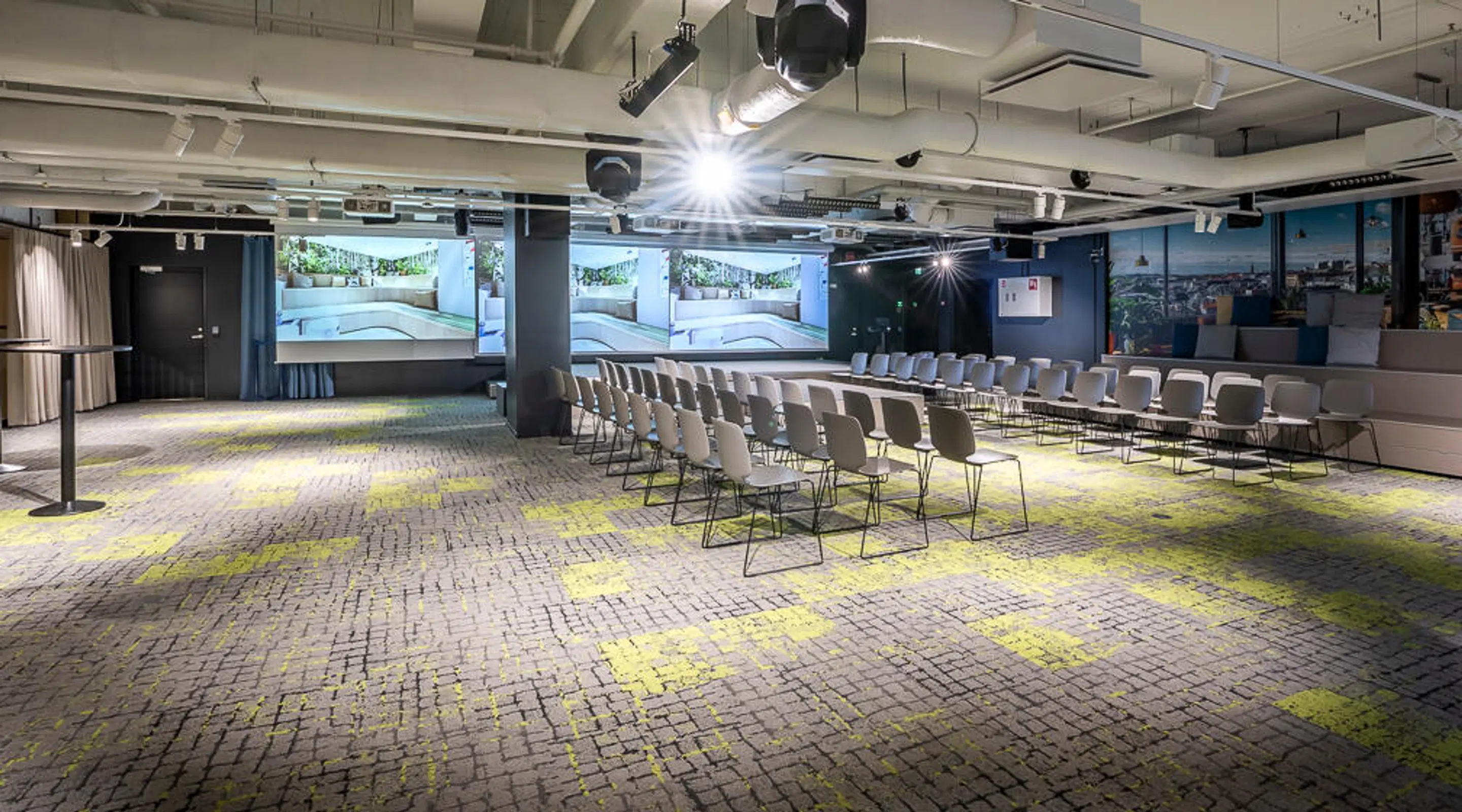 Modern auditorium med ljusa stolar, stora skärmar framtill, och mönstrad grå-gul heltäckningsmatta. Öppet takutrymme med teknik.