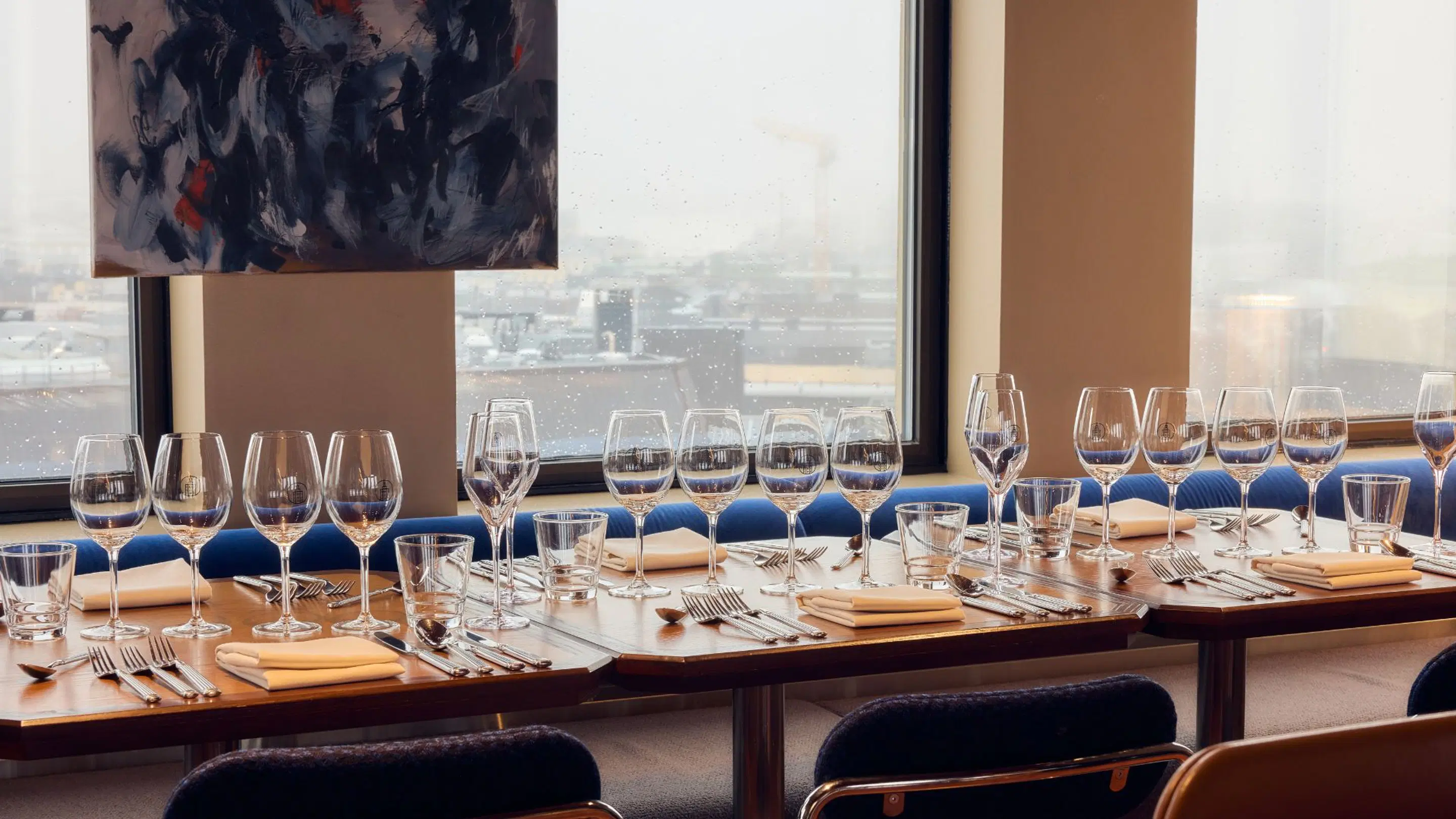 Restaurangbord dukat med vinglas och bestick. Stora fönster med stadsutsikt, mörk konst på väggen.