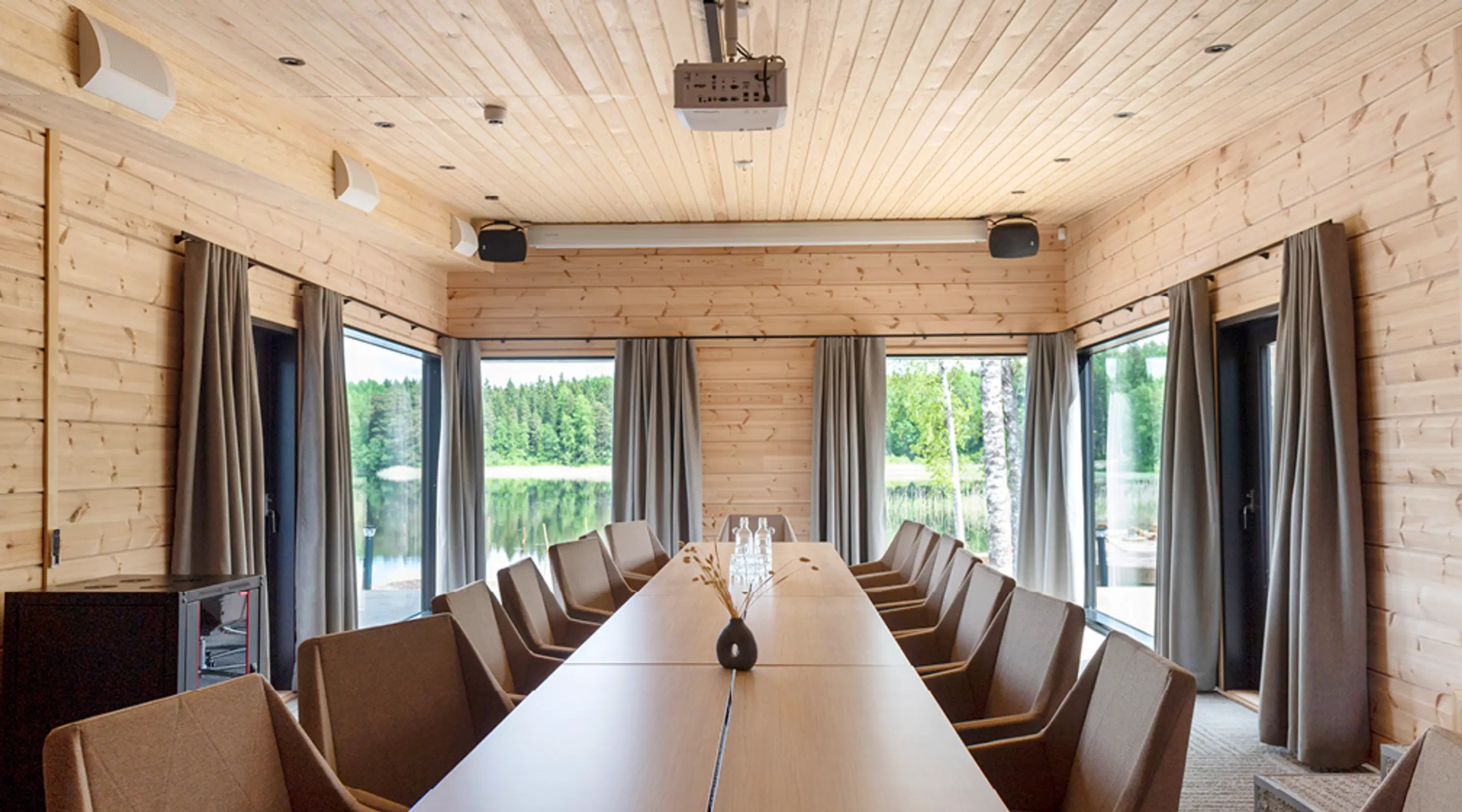Träpanelerat mötesrum med långt bord och stolar. Genom fönstren syns en sjöutsikt och skog. Projektor monterad i taket.