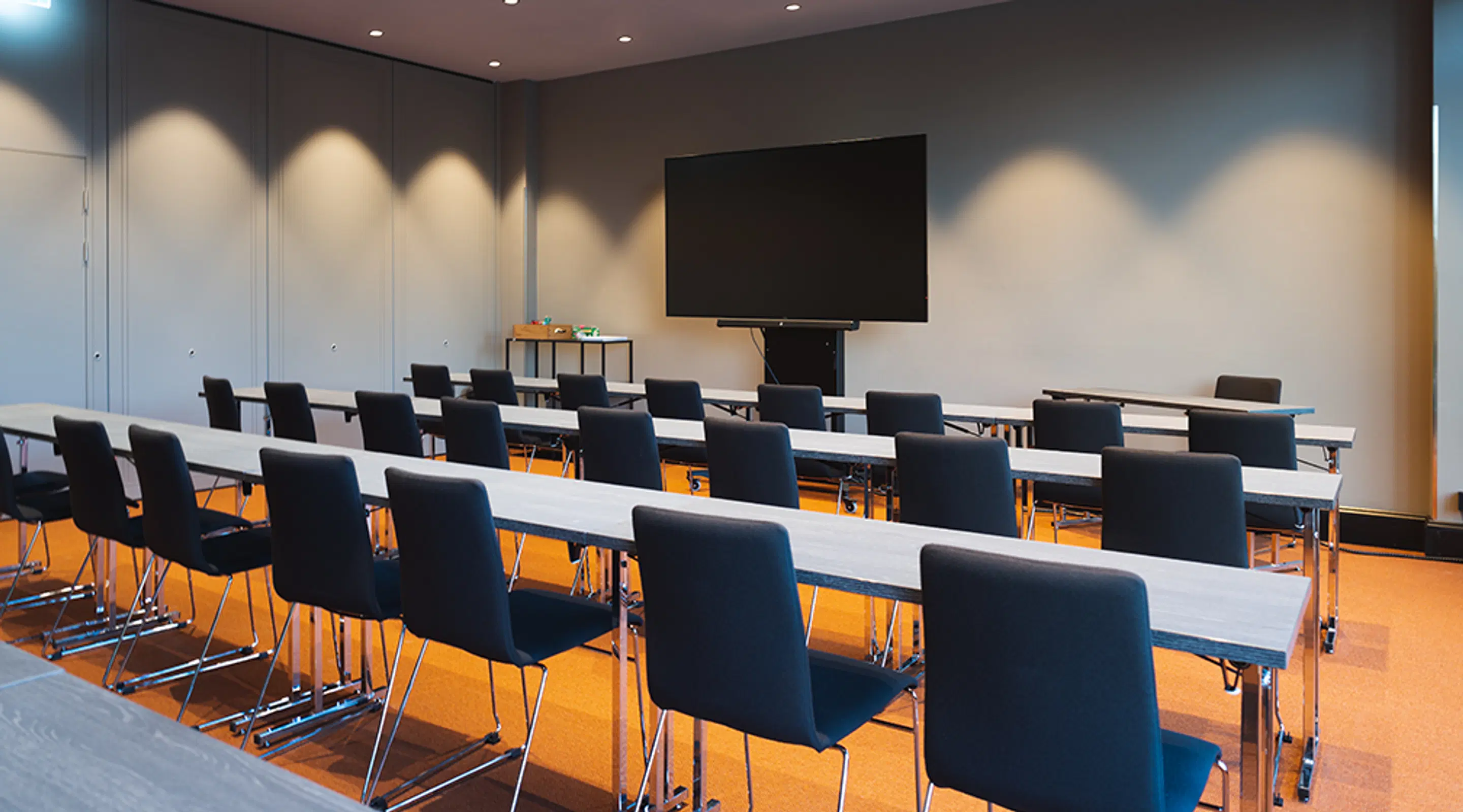 Modern conference room with rows of tables and blue chairs. Large display screen at front. Room has spotlights and orange carpeting.
