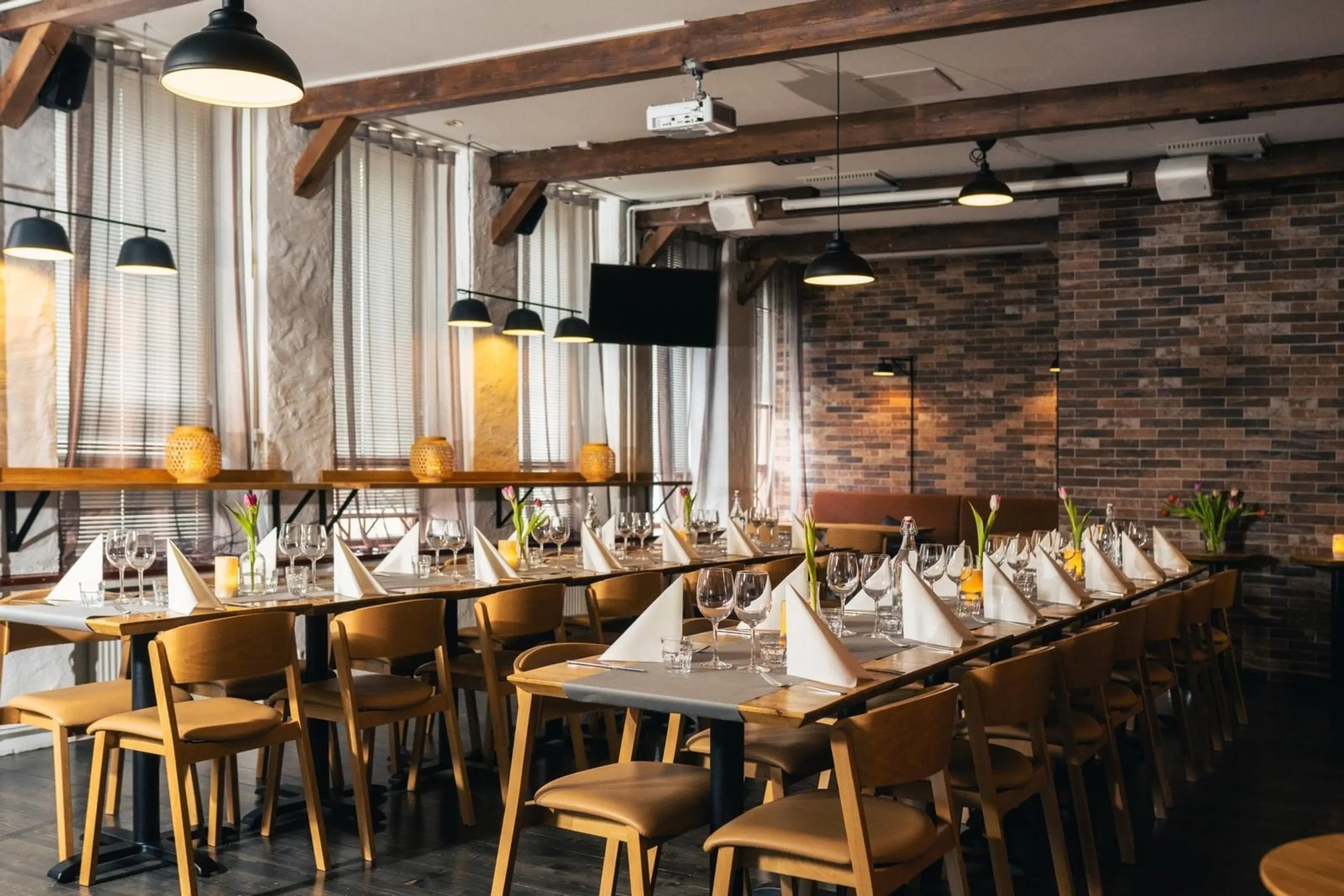 Restaurant interior with brick wall, wooden beams, and large windows. Tables set with glasses and napkins.