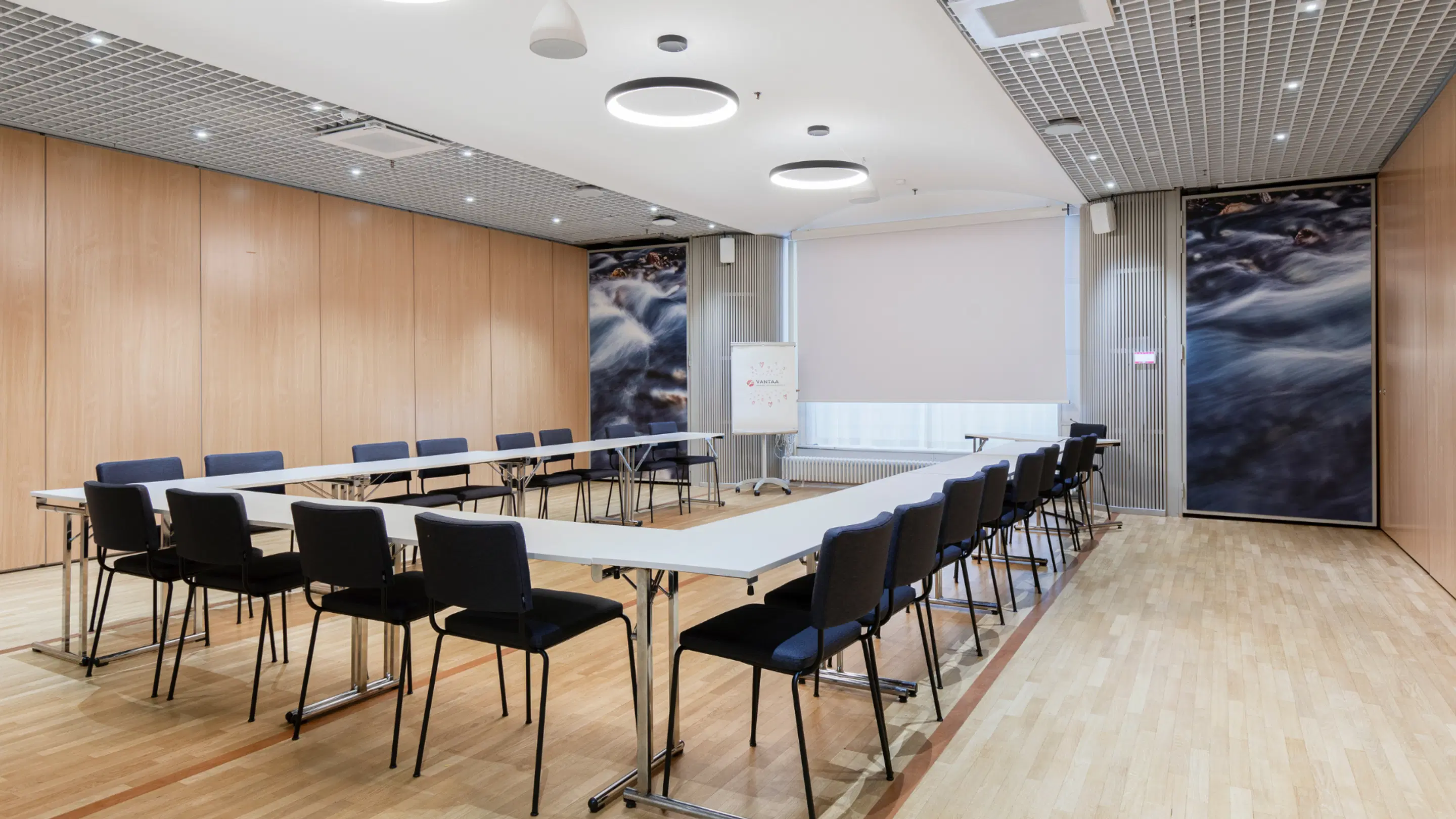 Modern meeting room with U-shaped table arrangement, wooden paneled walls and flowing water artwork. The room has even wooden flooring and good lighting.