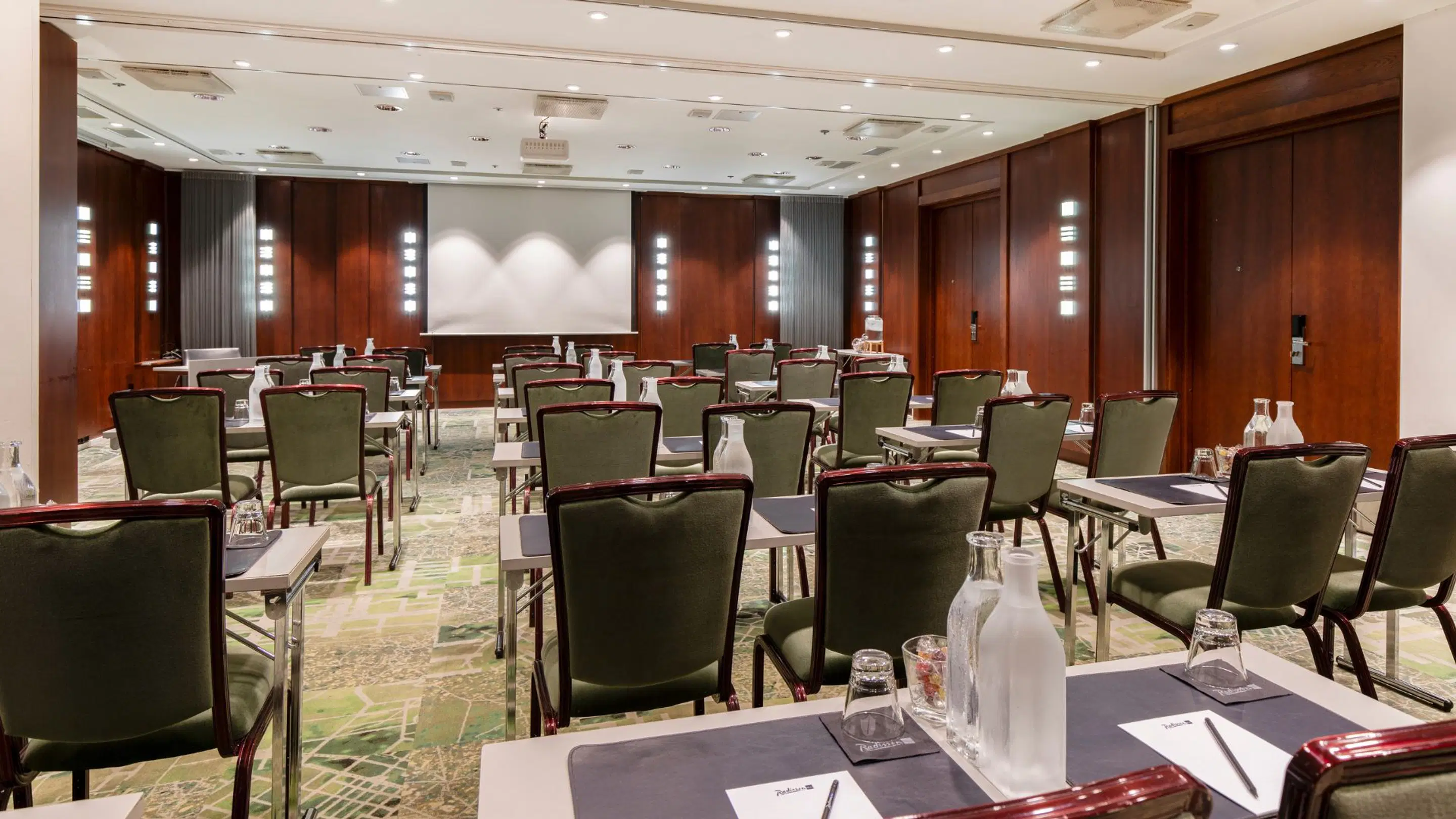 Conference room with wooden panel walls and white projector screen. Green chairs arranged in rows at tables. Even lighting and hard floor surface.