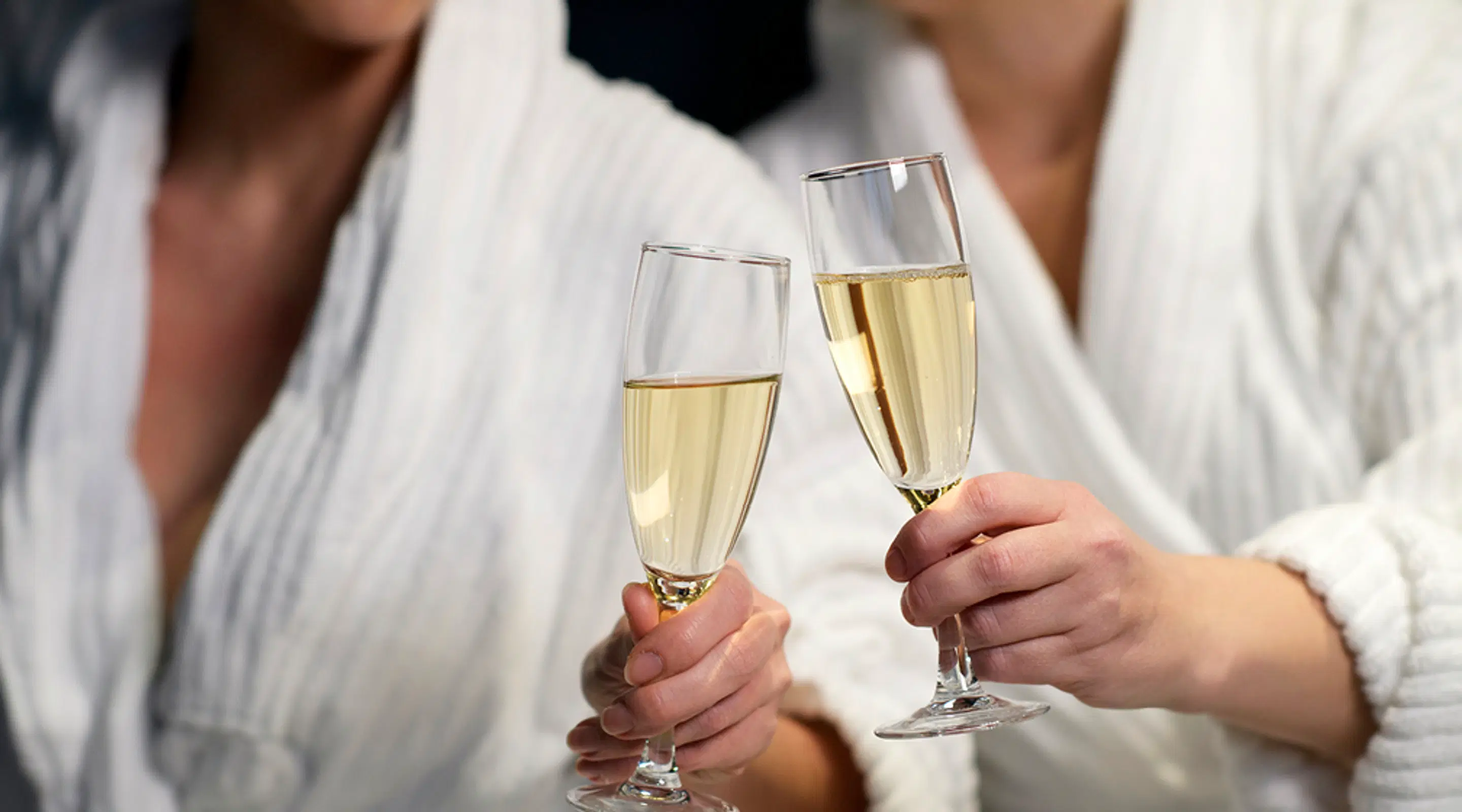 Two hands in bathrobes holding champagne flutes filled with sparkling beverage.