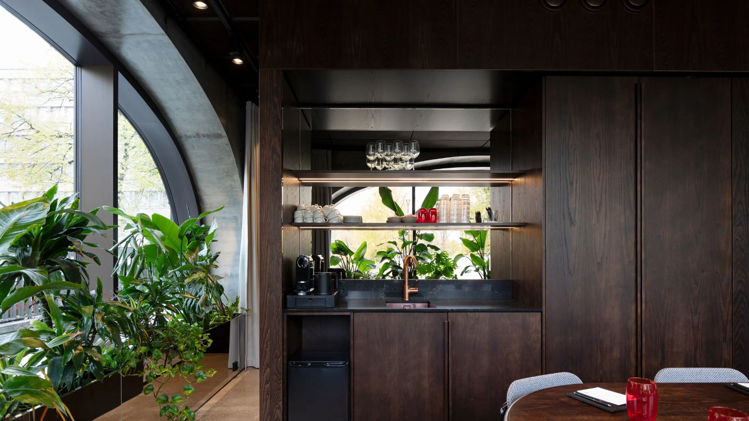 Modern compact kitchenette with dark wood paneling, lush plants by curved window, and small dining table.