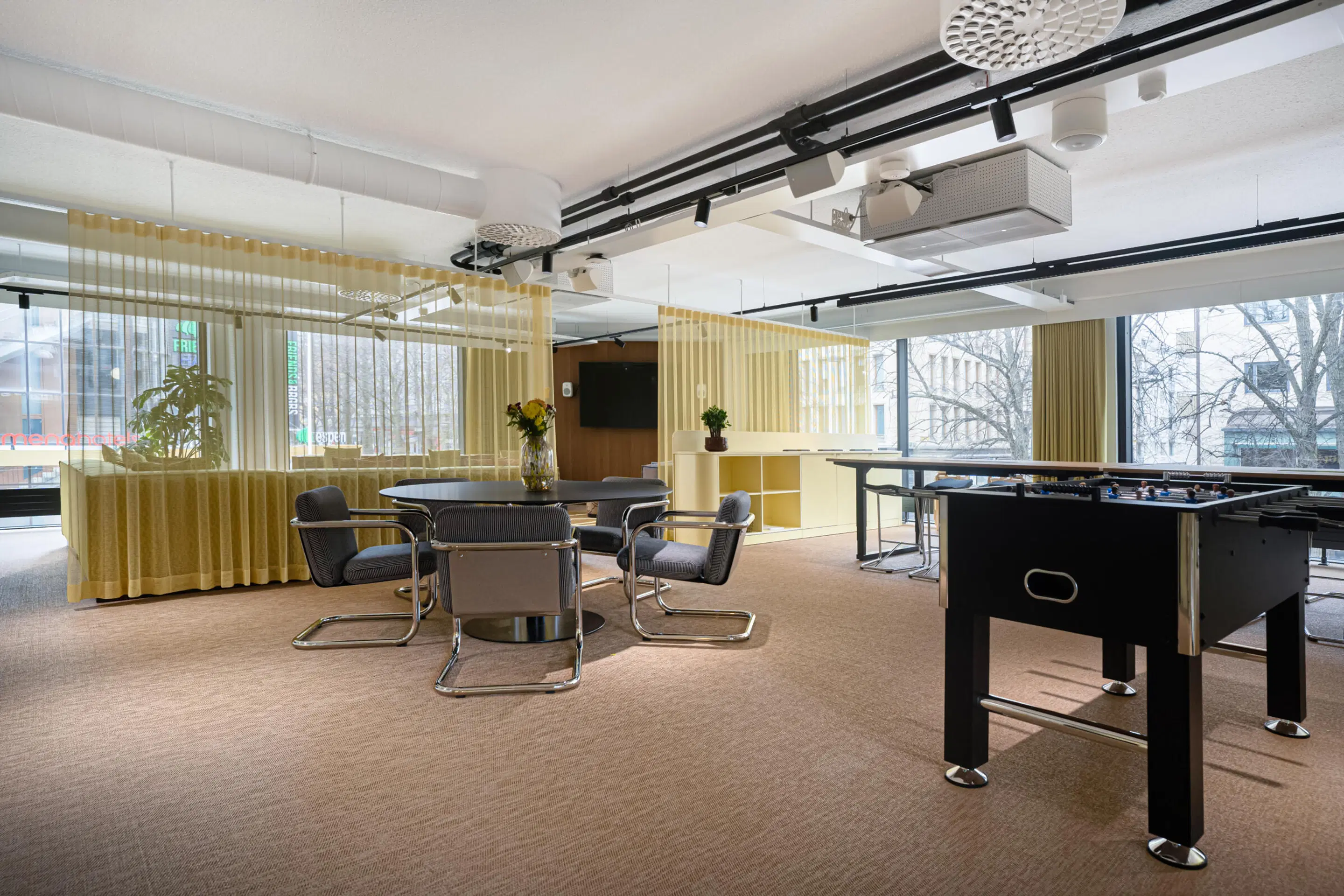 Modern break room with round table and chairs, foosball table, and yellow curtains. Level carpet flooring and large windows.