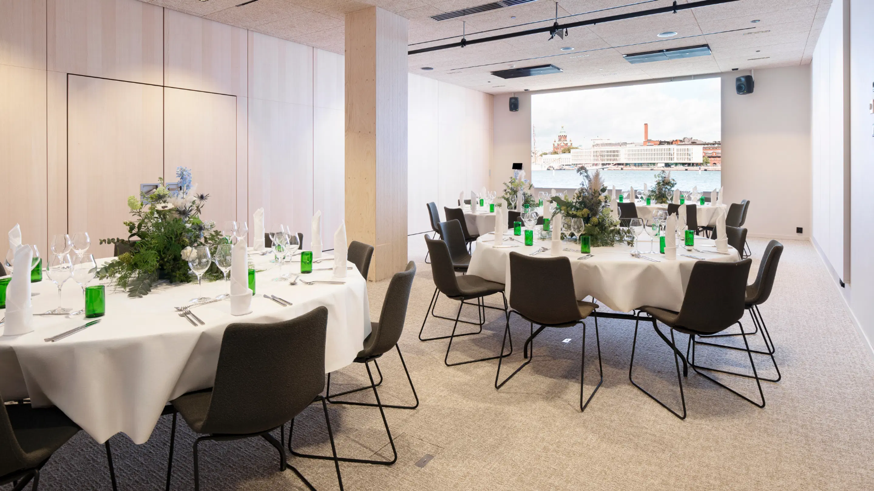 Spacious banquet room with round tables, white tablecloths and floral arrangements. Large screen with city view in background and level carpeted flooring.