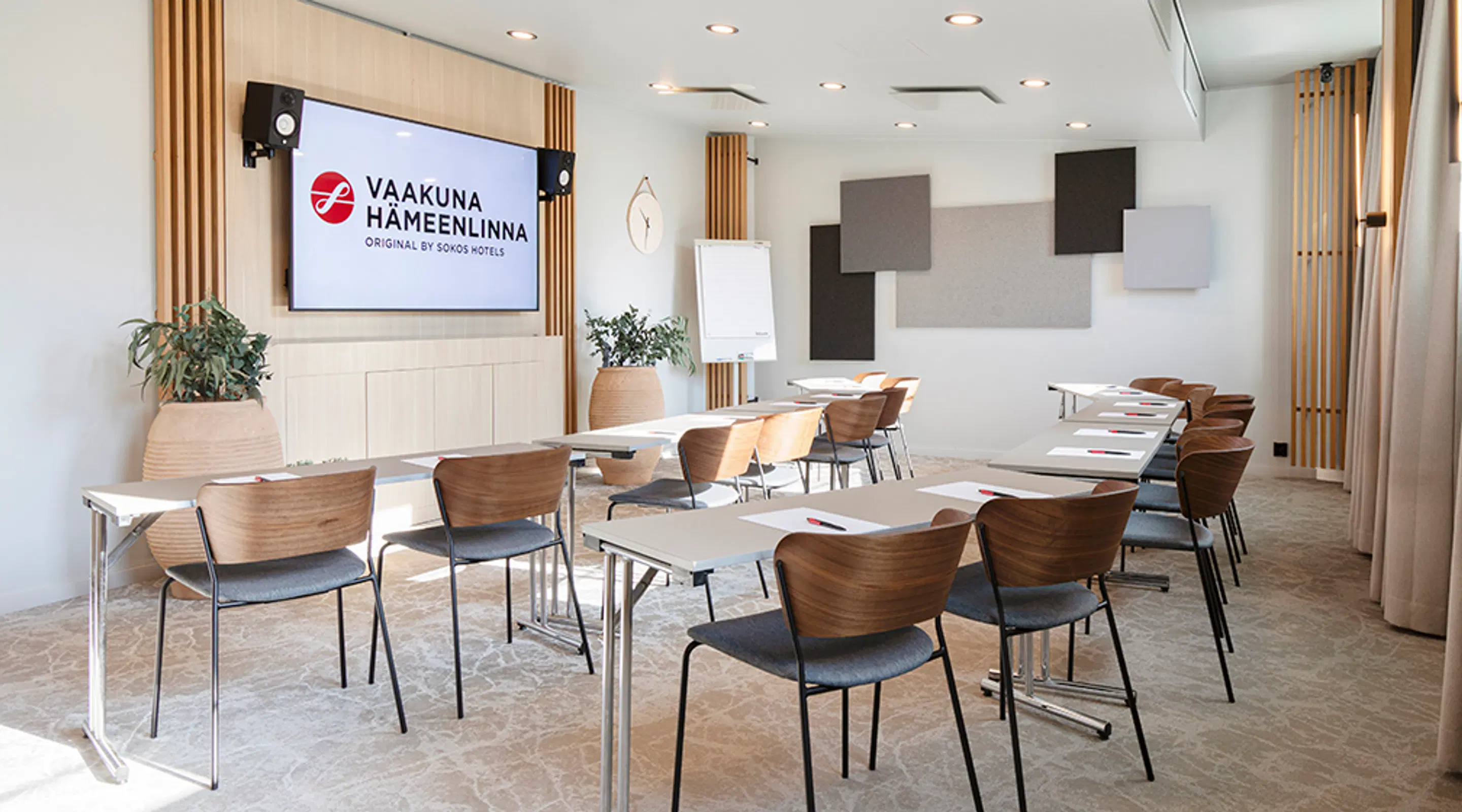 Moderni kokoustila hotellissa, jossa pöytiä ja puisia tuoleja. Suuri näyttö seinällä, akustiikkapaneeleita ja viherkasvi. Tilava lattia ja hyvä valaistus.
