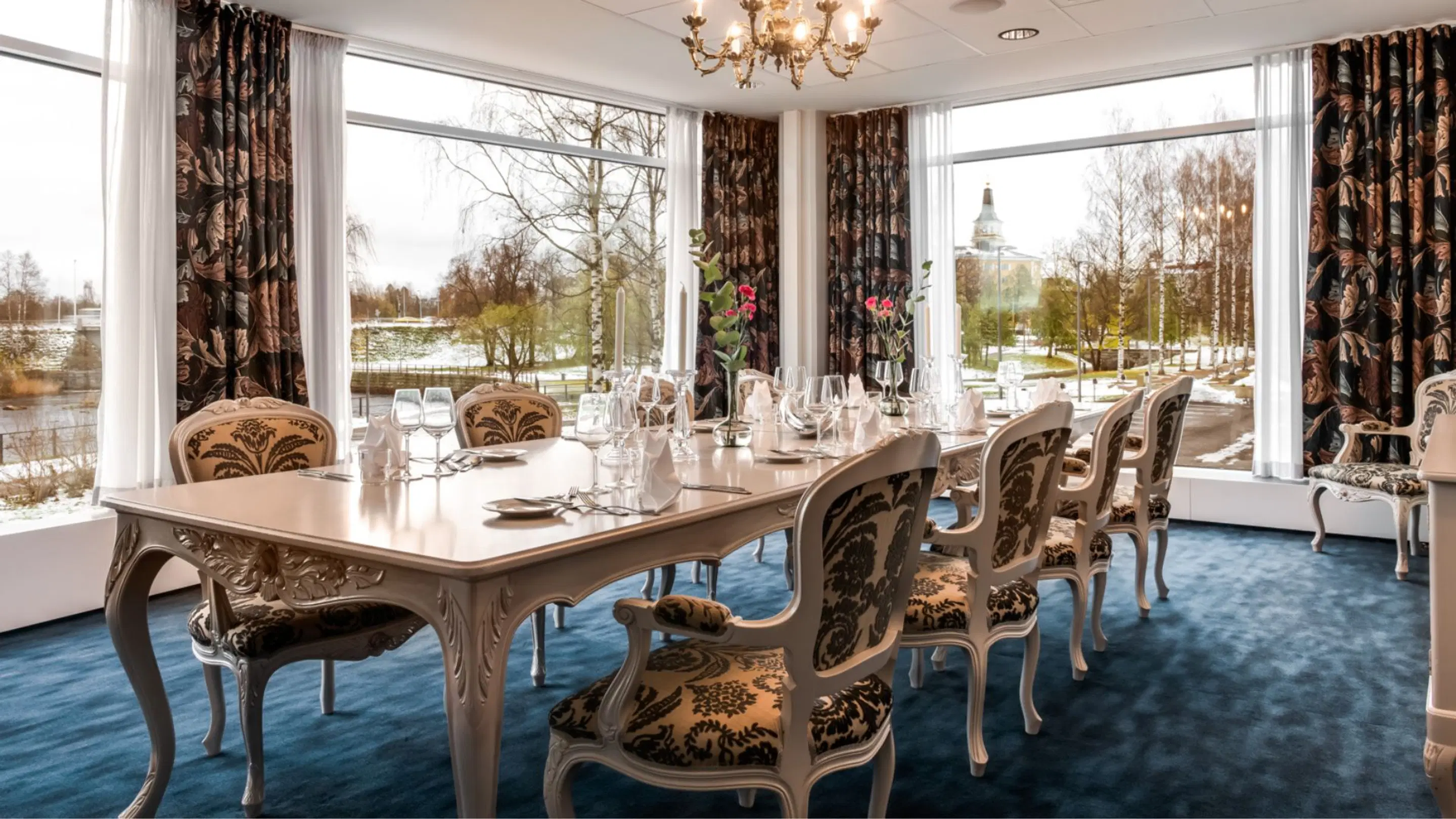 Elegant dining room with white table and upholstered chairs, blue carpet and large windows overlooking winter landscape. Chandelier illuminates the space.