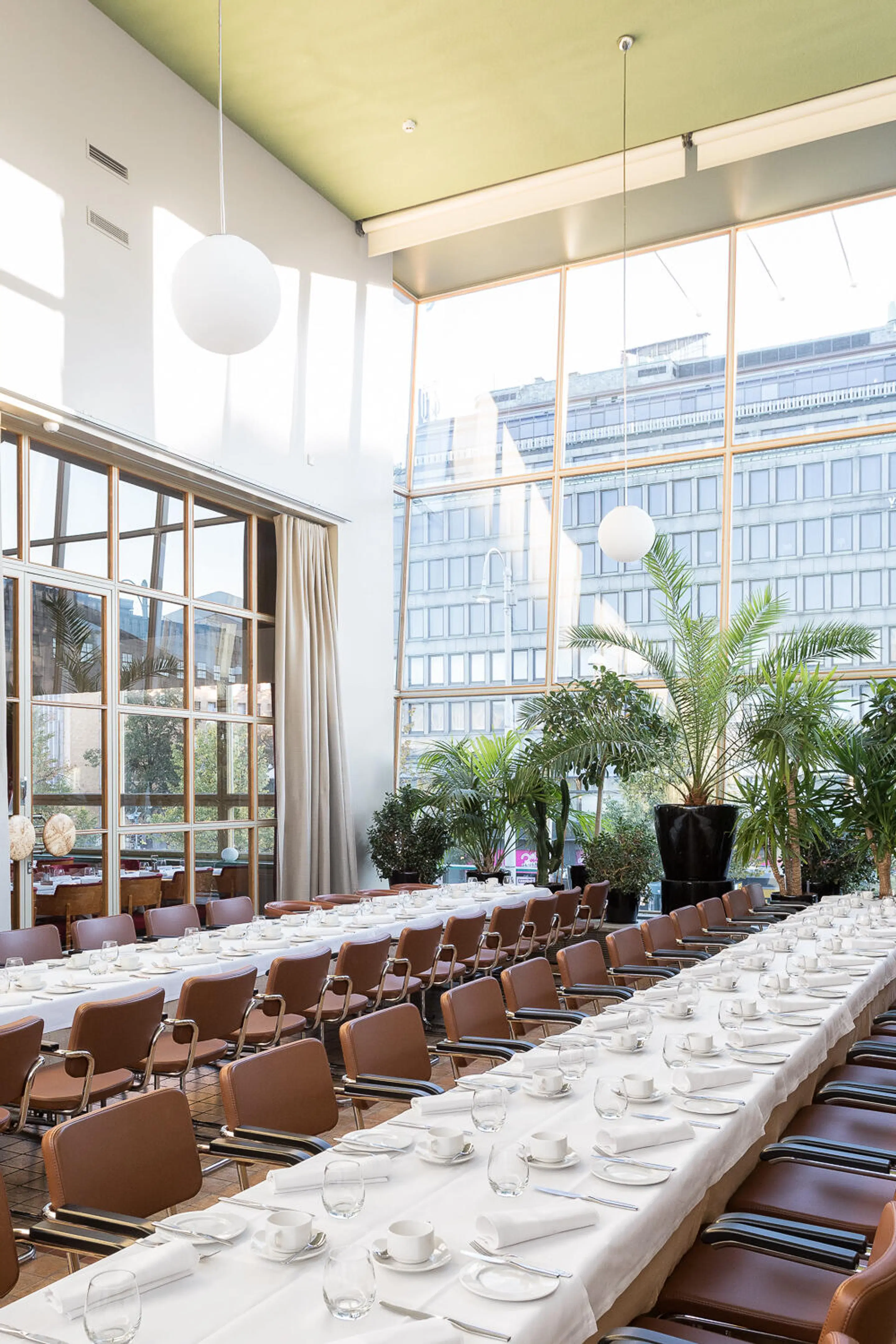 Bright restaurant dining hall with long tables covered in white tablecloths, brown chairs, and abundant greenery. Tall windows offer city views.