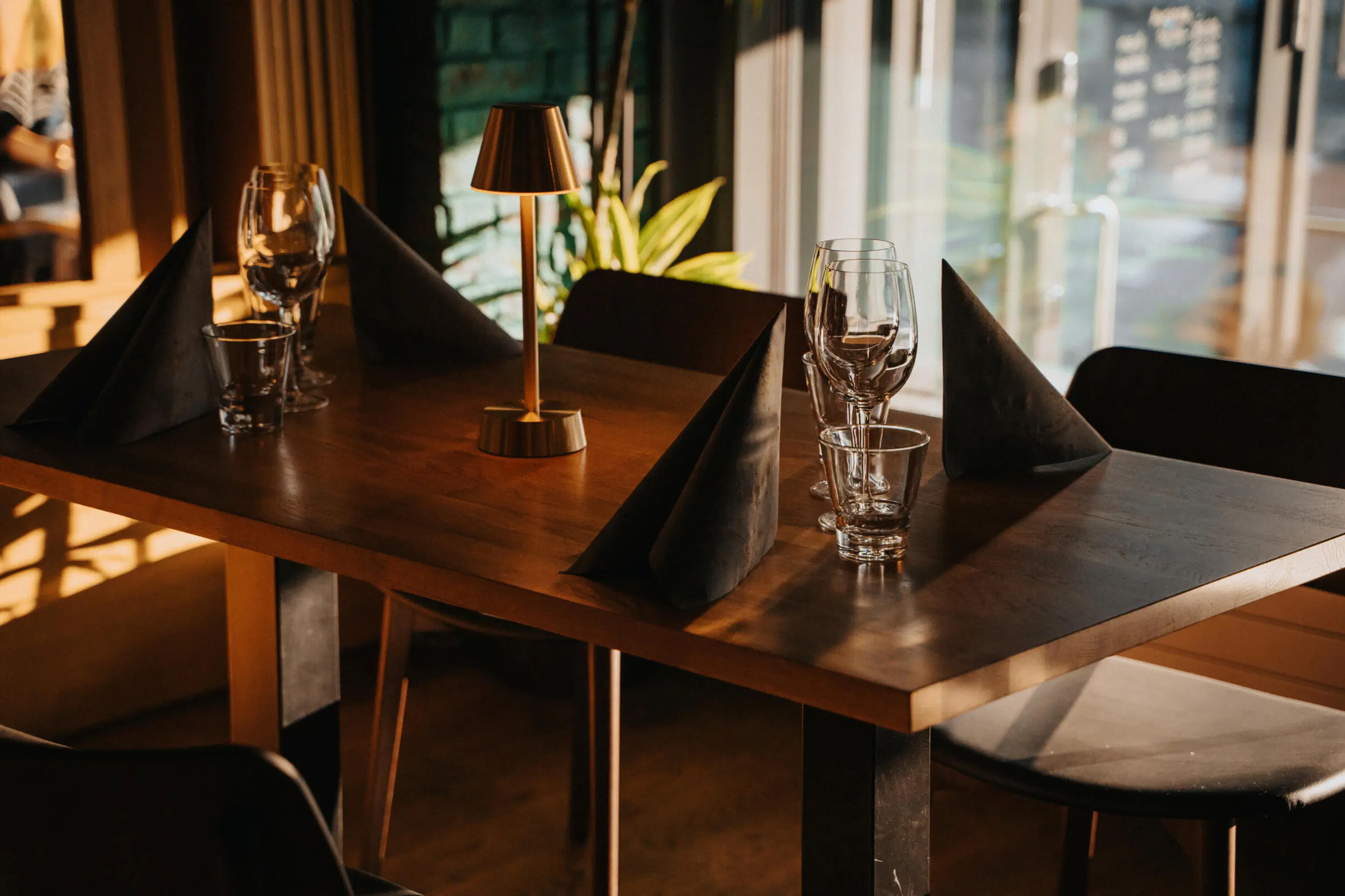 Elegantly set restaurant table with wine glasses, black napkins and a gold table lamp. Large windows beside the table.