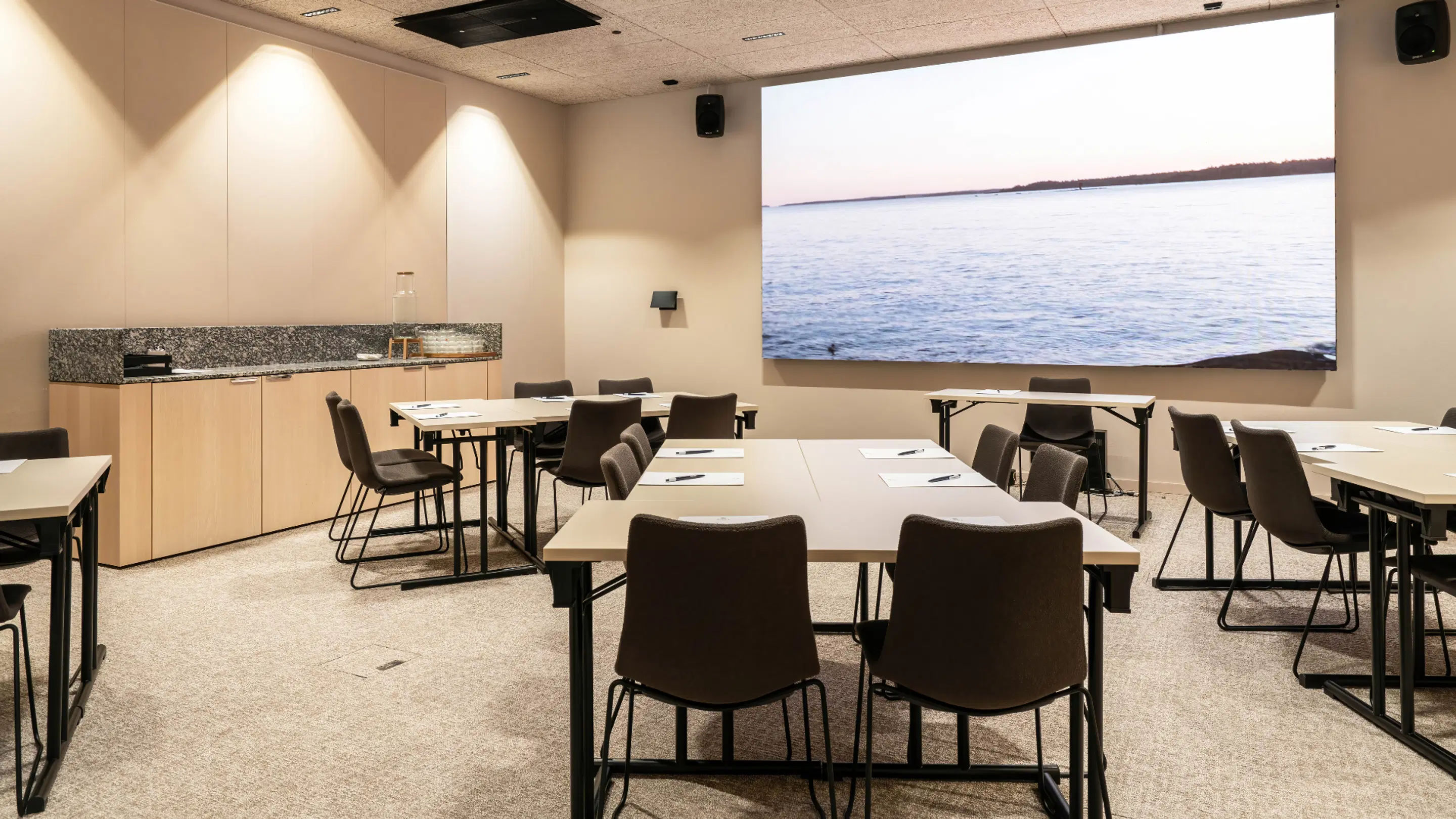 Conference room with table arrangements and a large screen displaying ocean view. Space includes small serving area with granite countertop.