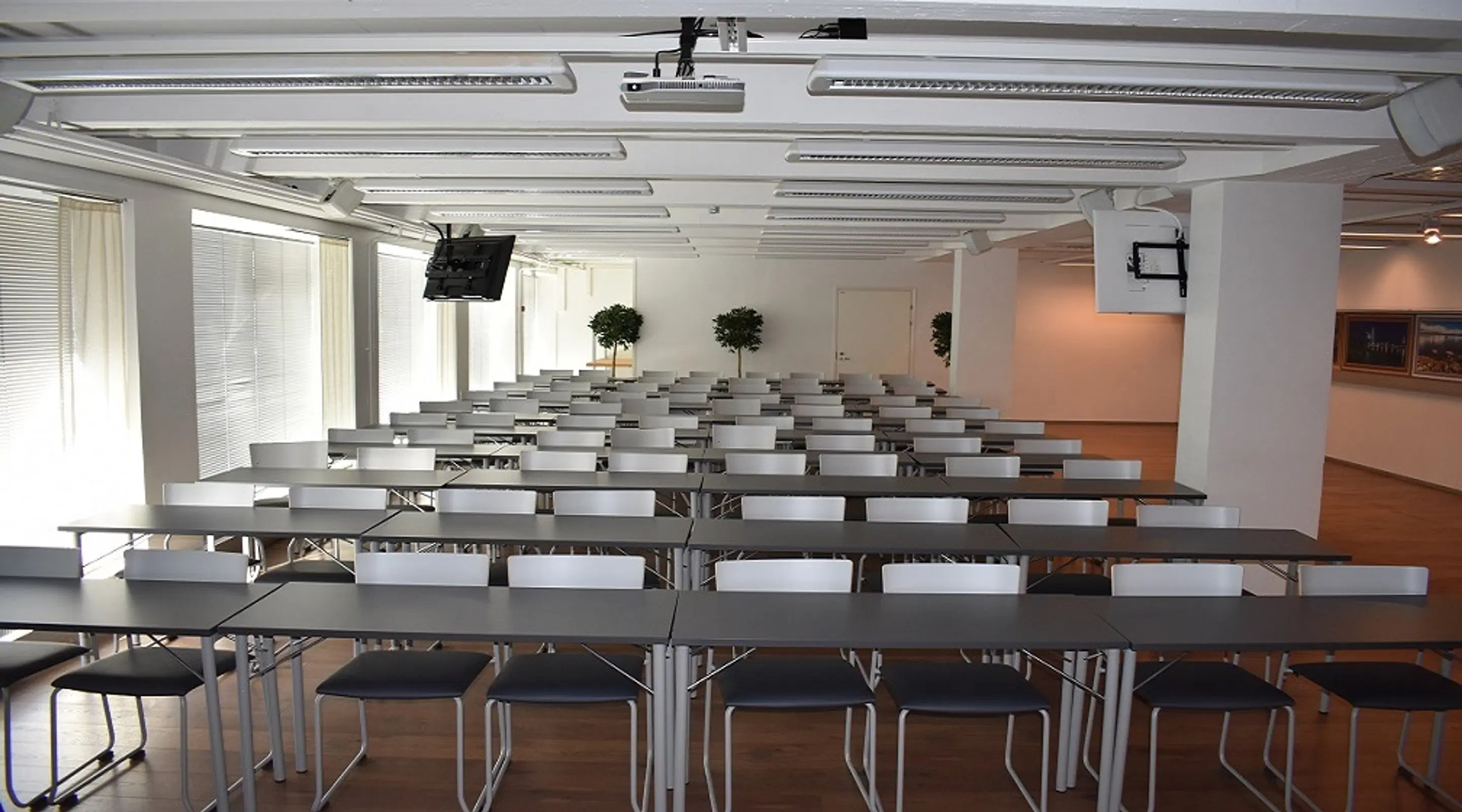 Conference room with rows of tables and chairs, level wooden floor, white walls and ceiling with rows of lights. Small decorative plants are also present.