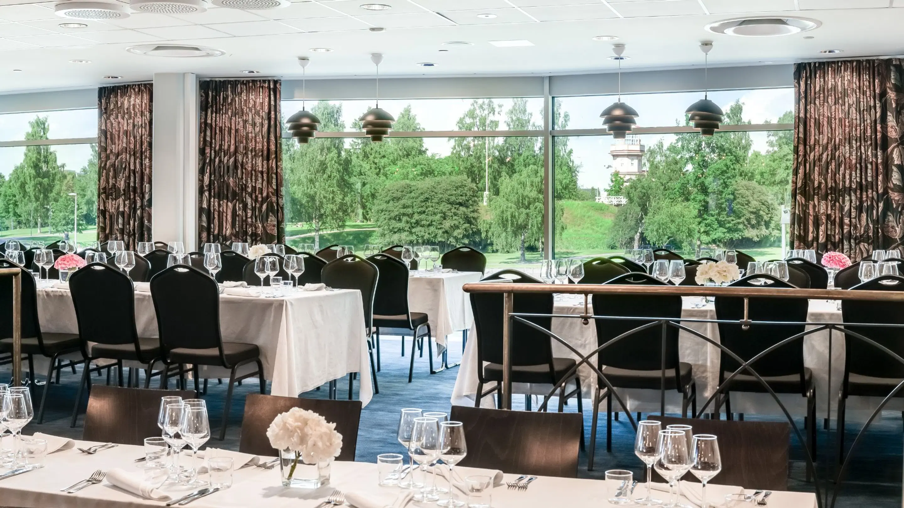 Banquet hall with tables and chairs. Large windows overlook a lush green park. Level floor with ample space for movement between tables.