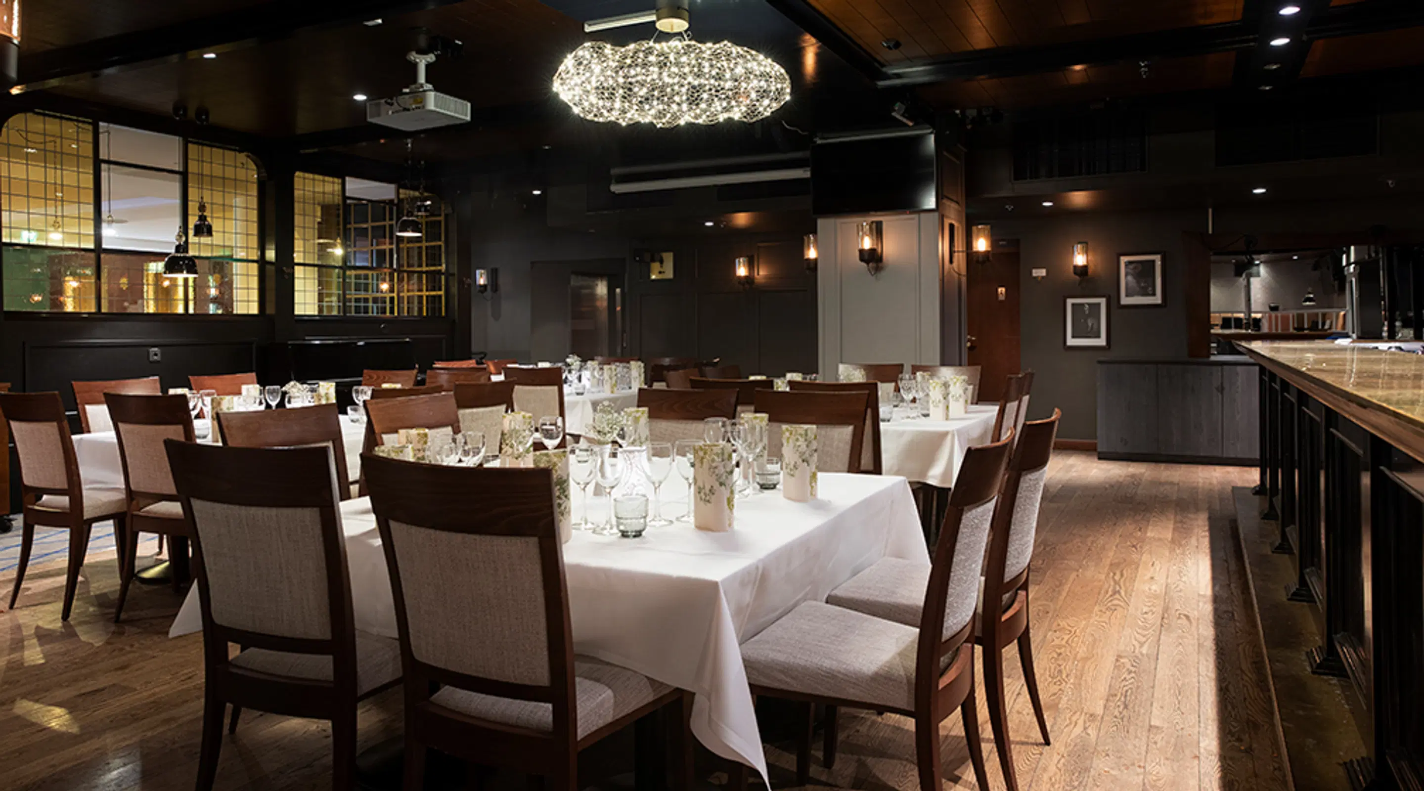 Elegantne restoranisaal puitpõranda, tumedate seintega ja valgete laudlinadega kaetud laudadega. Lühtriga valgustatud ruum.
