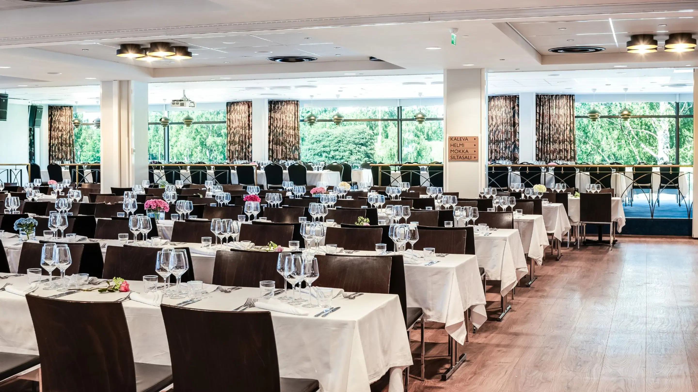 Bright restaurant dining hall with dark chairs and white tablecloths. Wine glasses and small flower arrangements on tables. Large windows overlook lush greenery.