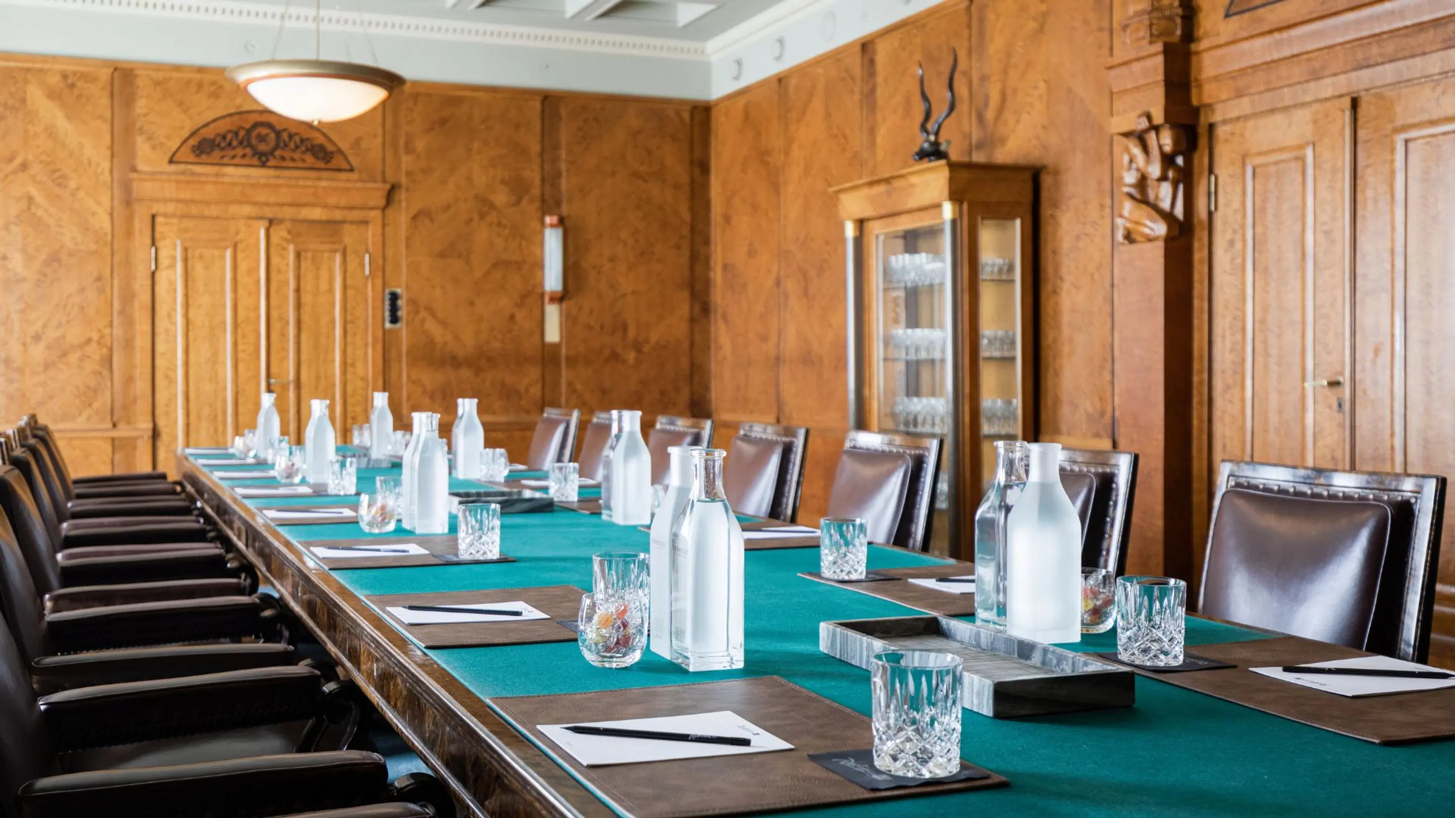 Meeting room with wooden wall panels, long table with turquoise-green tablecloth, leather chairs and glass bottles on the table.