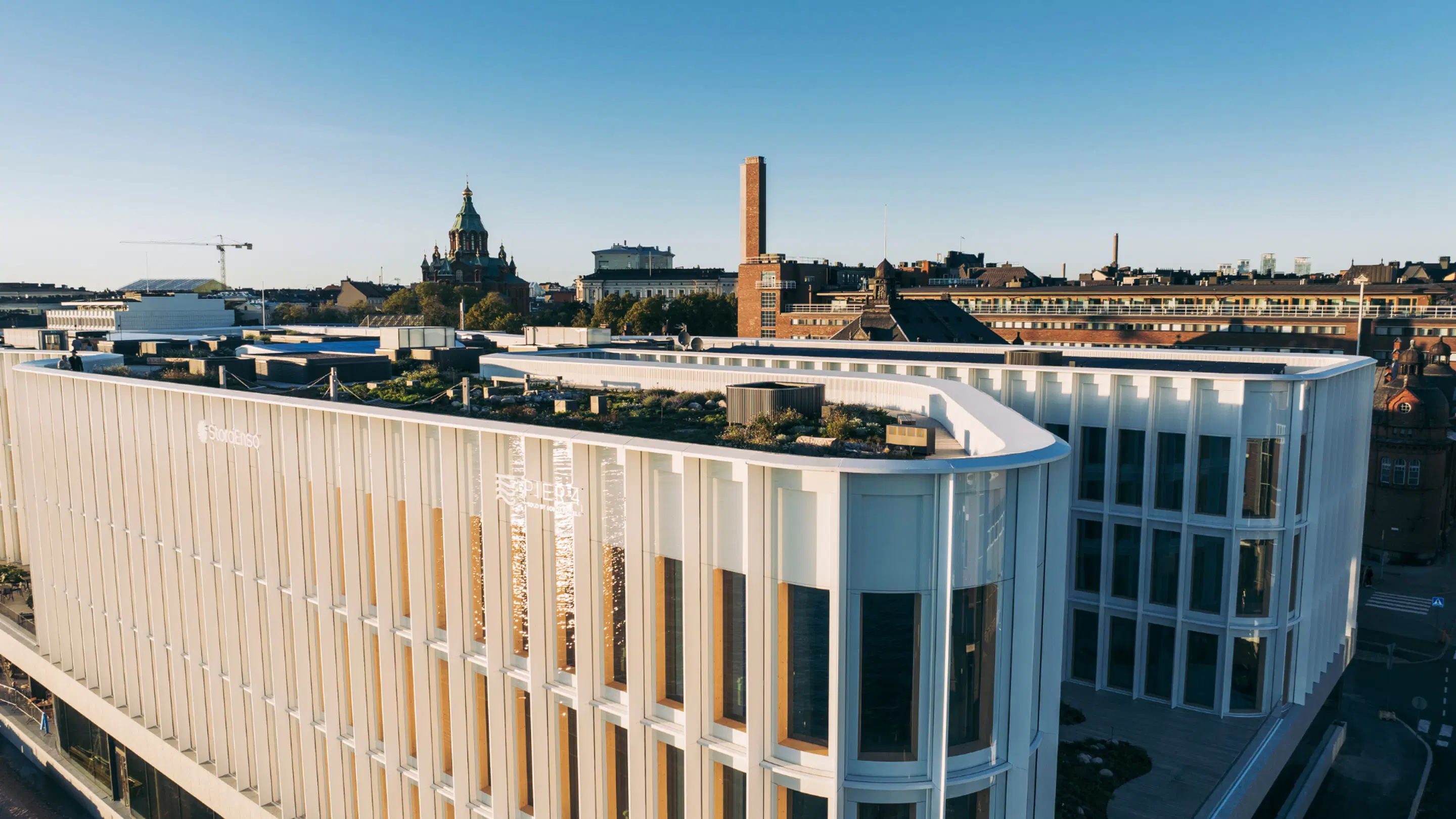 Modern white building with green roof garden, with Uspenski Cathedral and Helsinki cityscape visible in the background.