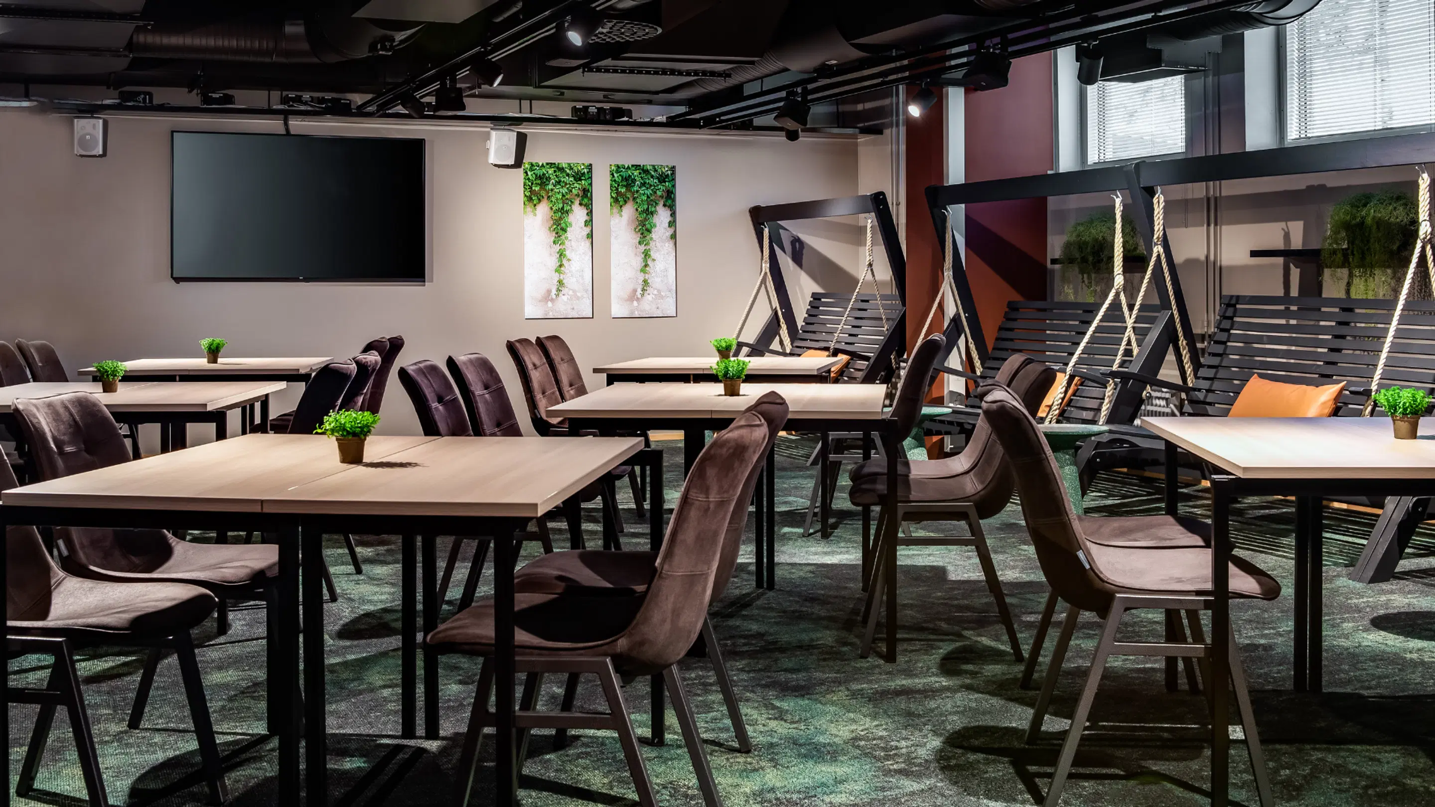 Modern meeting space with table arrangements, display screen and swing seats. Green plants on tables and wall art. Level flooring and ample space to move around.
