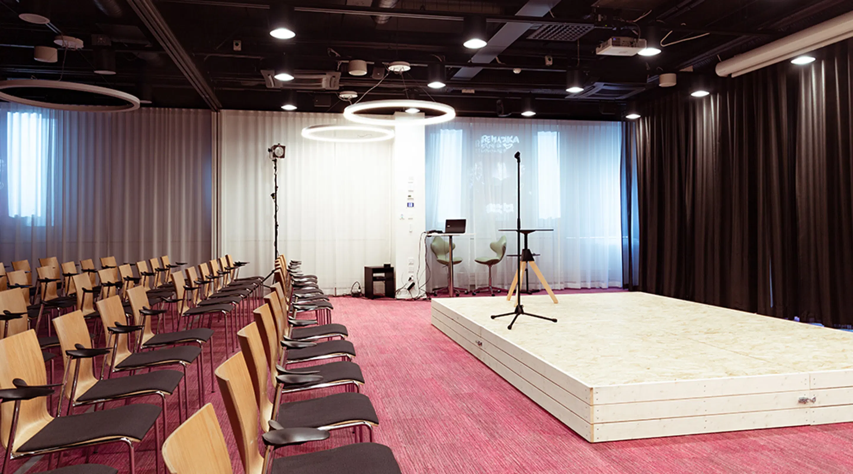 Modern auditorium with light wooden stage and red carpet. Rows of chairs with wooden backrests and microphone in front of the stage.