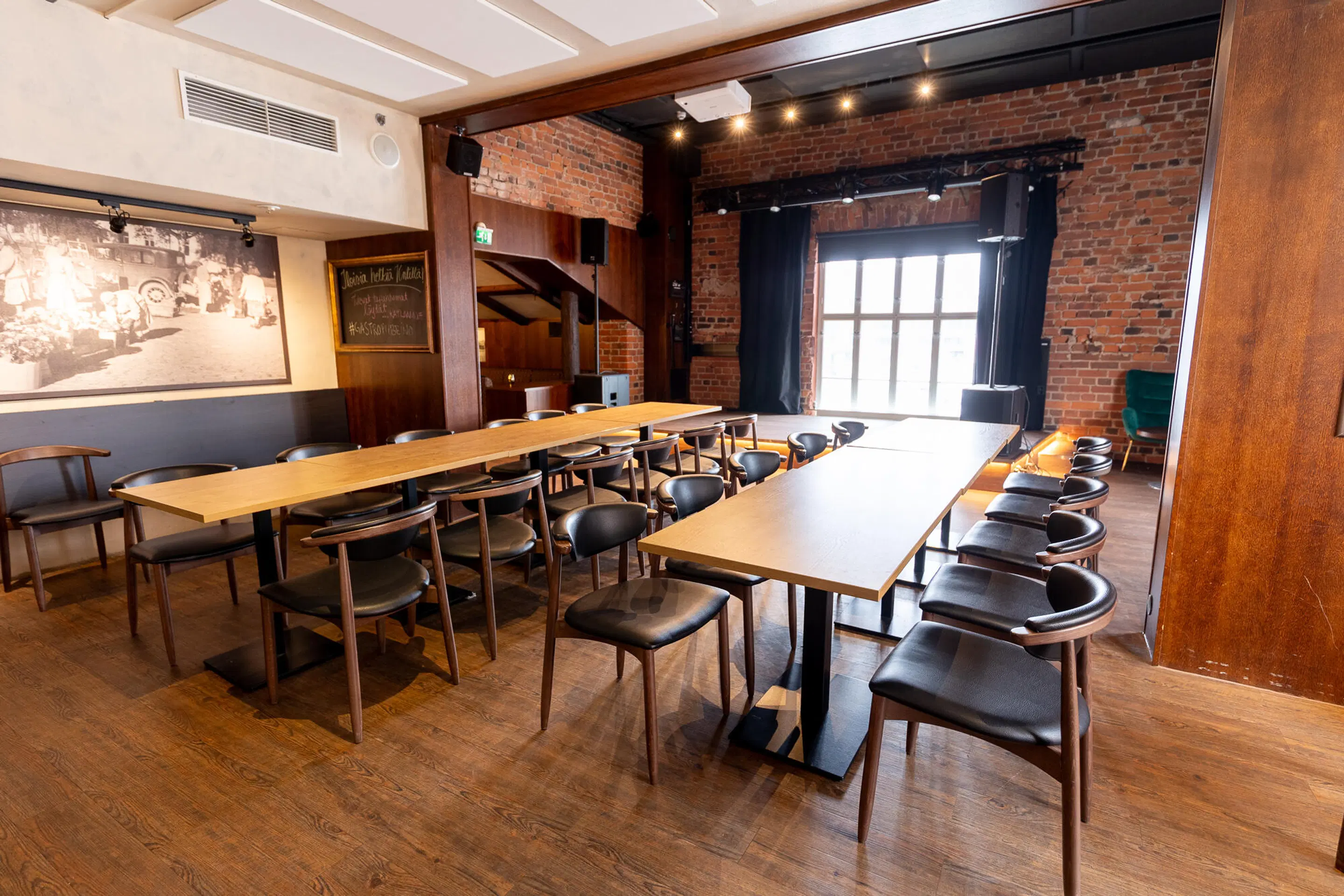 Meeting room with two long tables and chairs on wooden flooring. Brick walls, large window and a small stage area in the background. Hard floor surface.