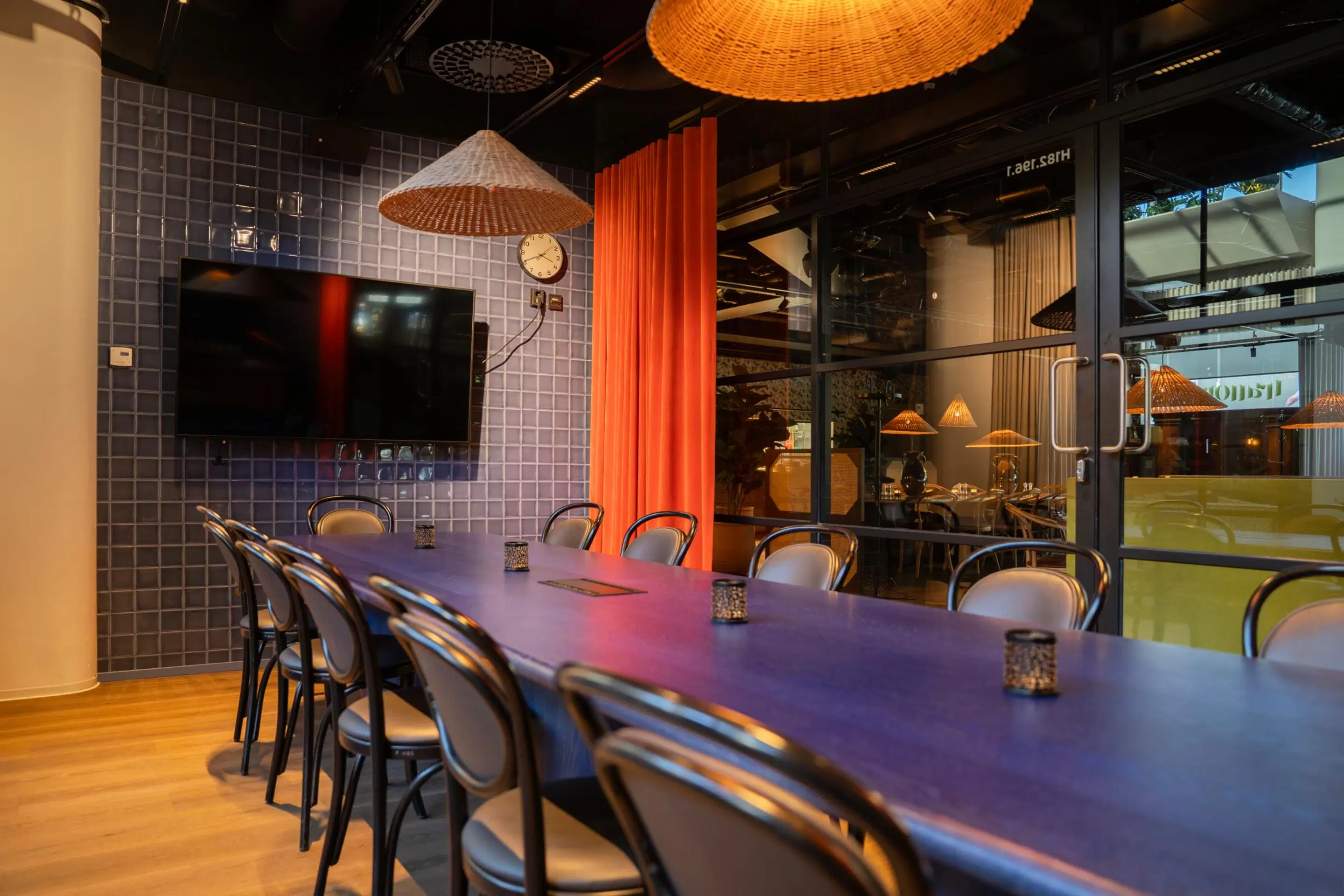 Modern meeting room with a long table, wicker pendant lights, and blue tiled wall. Space features wooden flooring and glass partition.