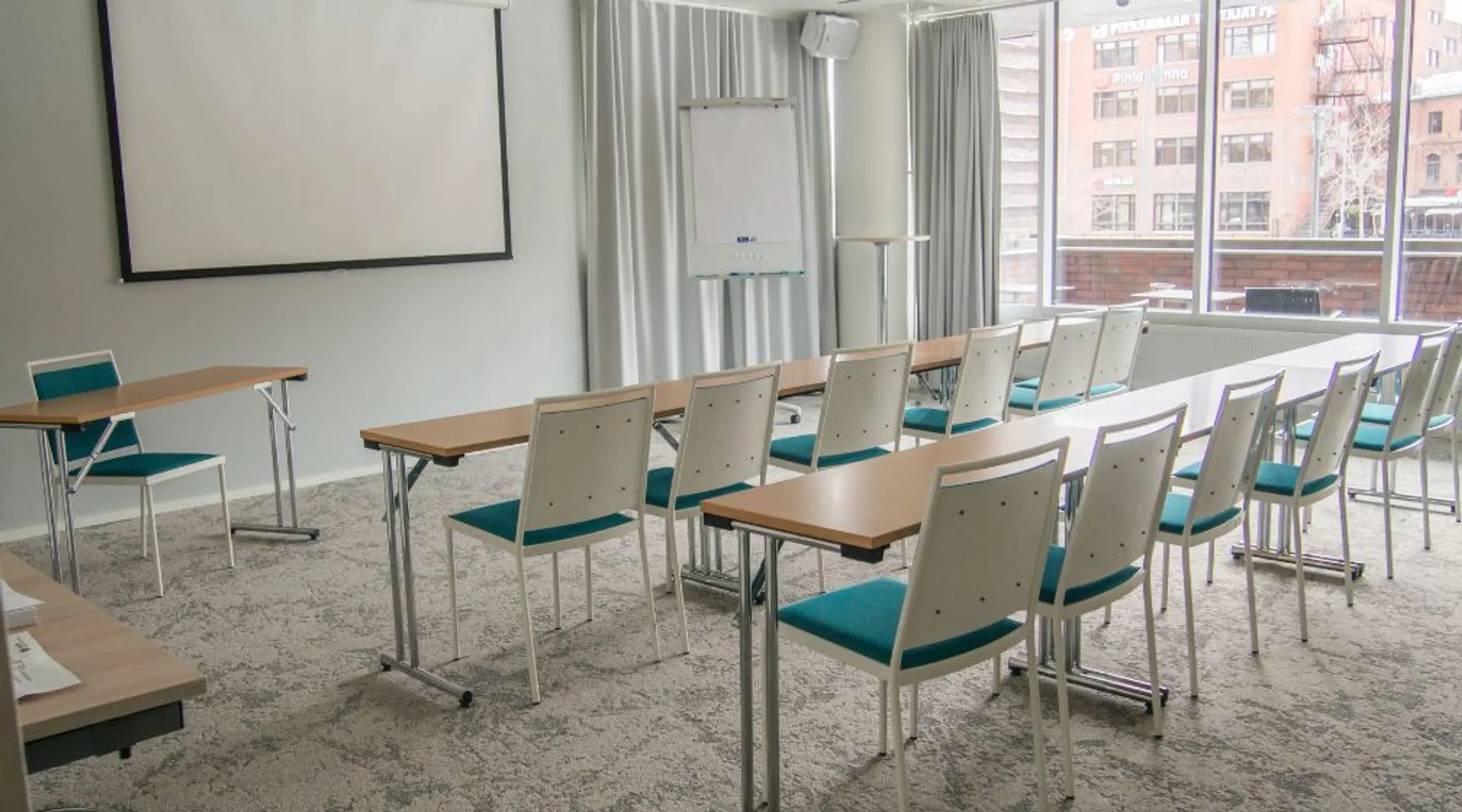 Modern meeting room with rows of lightweight tables, light-colored chairs with teal seats, a projection screen, and large windows overlooking city buildings.