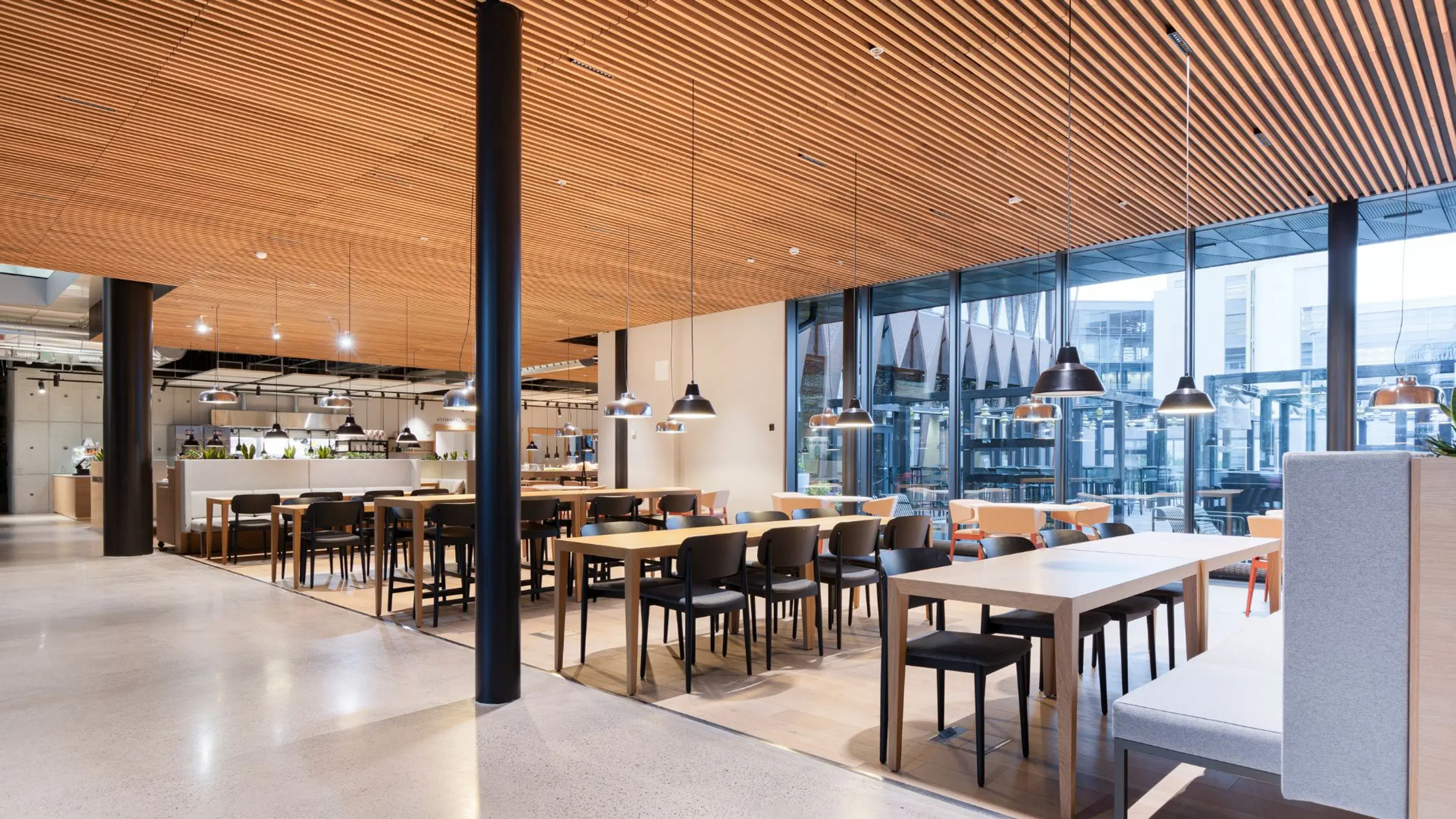 Modern cafeteria with wooden ceiling paneling, open floor plan and large windows. Smooth floor and wide pathways between tables.