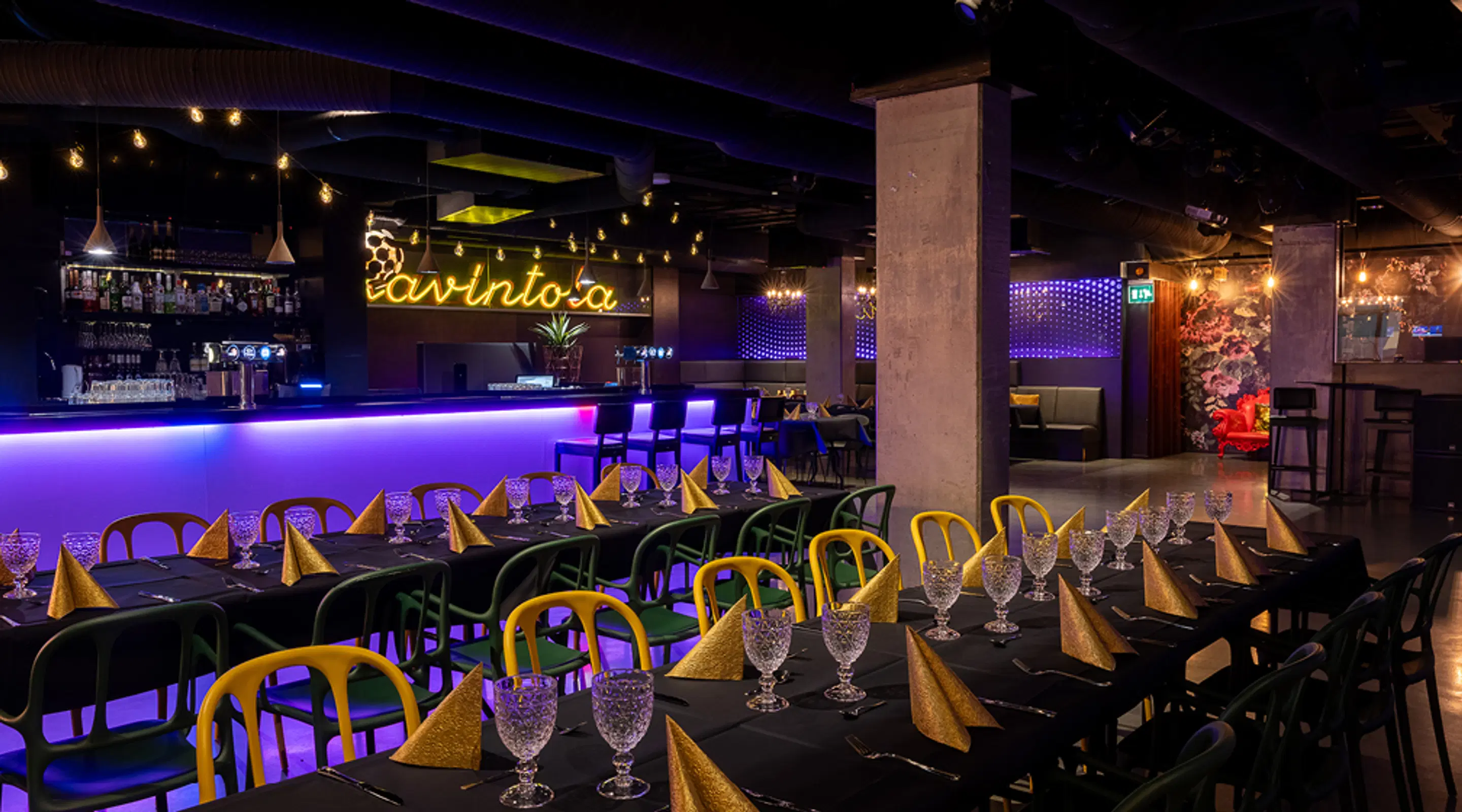 Atmospheric restaurant with dark tables, colorful chairs, crystal glasses and gold napkins. Space illuminated by purple light bar and hanging string lights.