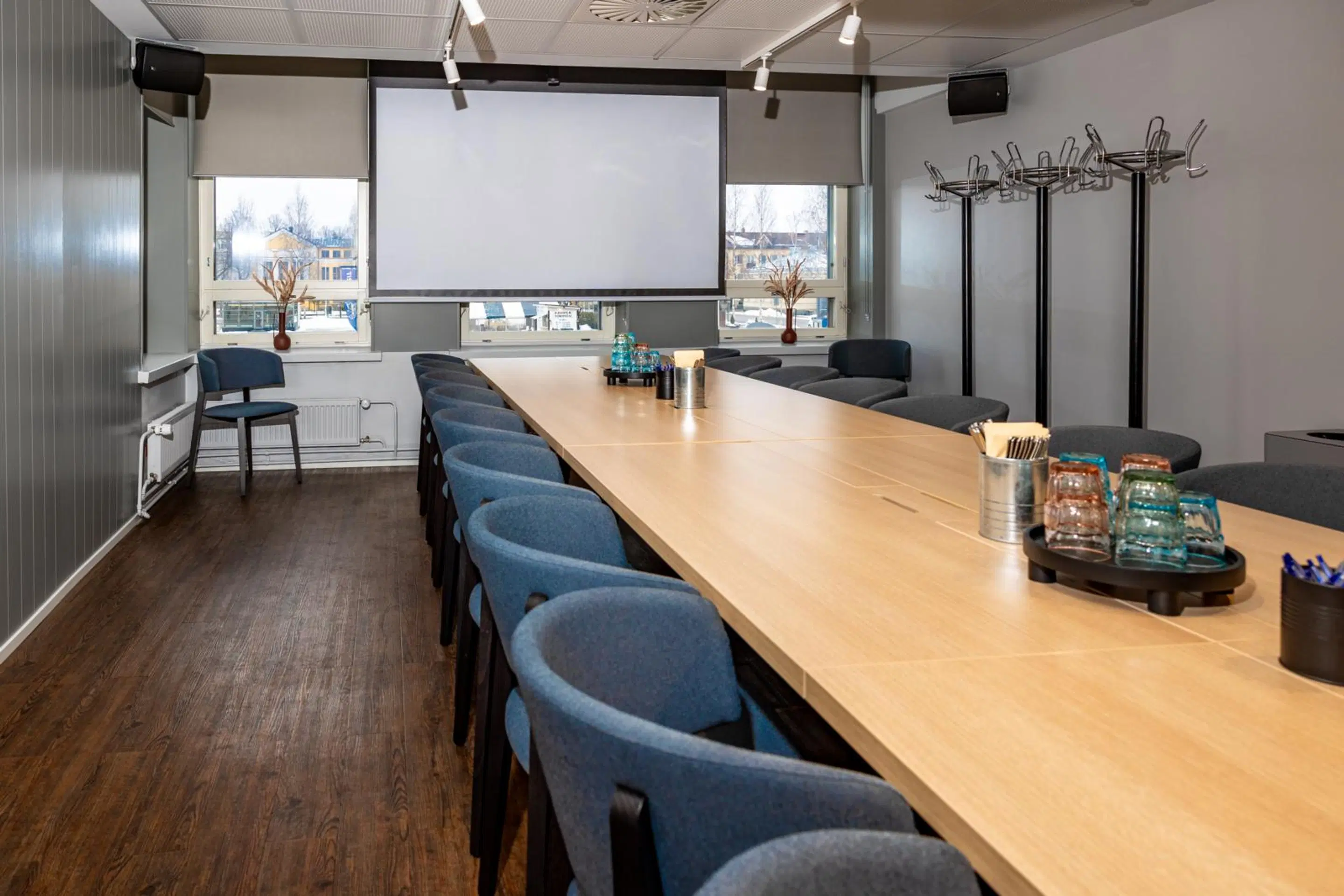 Conference room with long wooden table, blue chairs, projection screen and coat rack. Wooden floor and large windows.