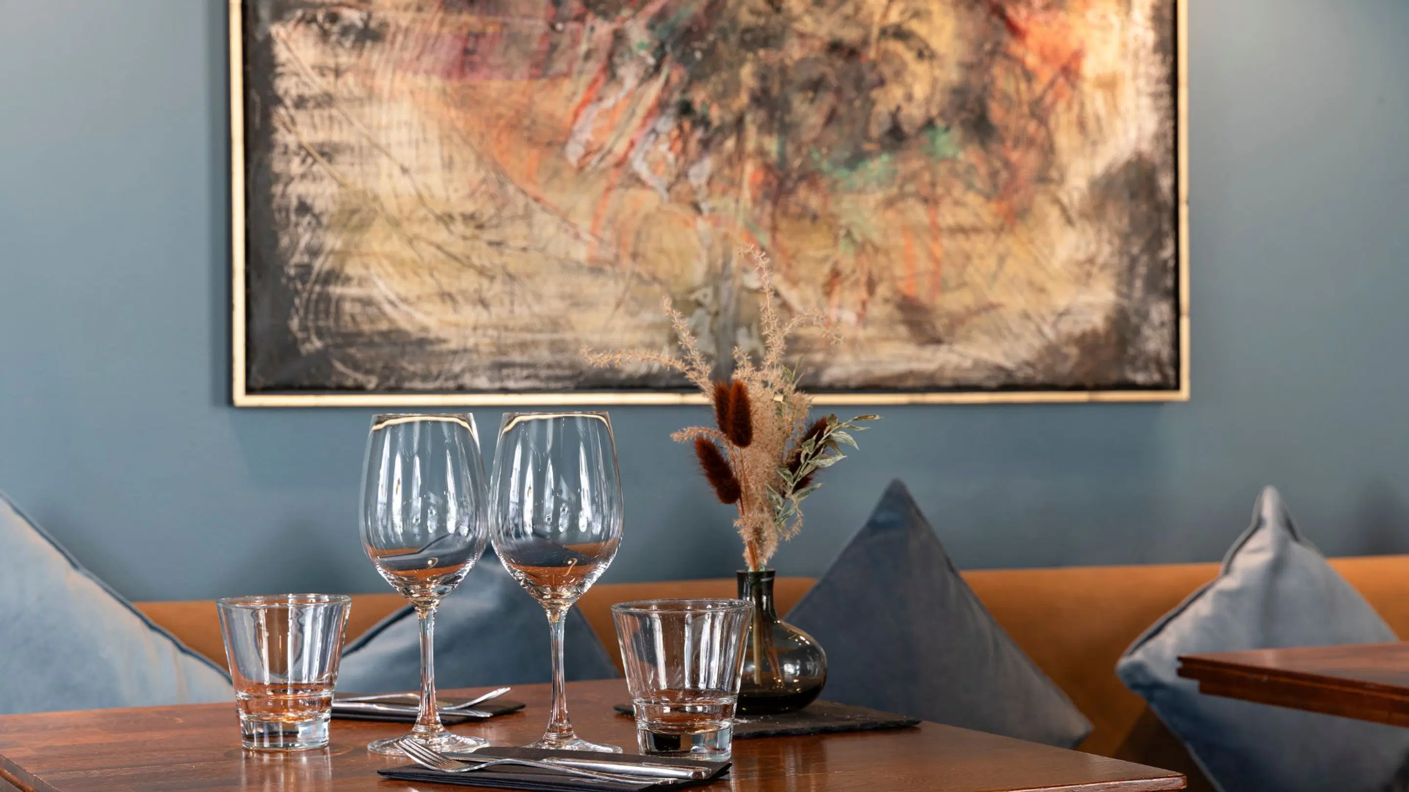Restaurant table with two wine glasses, drinking glasses, cutlery and a small vase of dried flowers. Abstract artwork on blue wall behind.