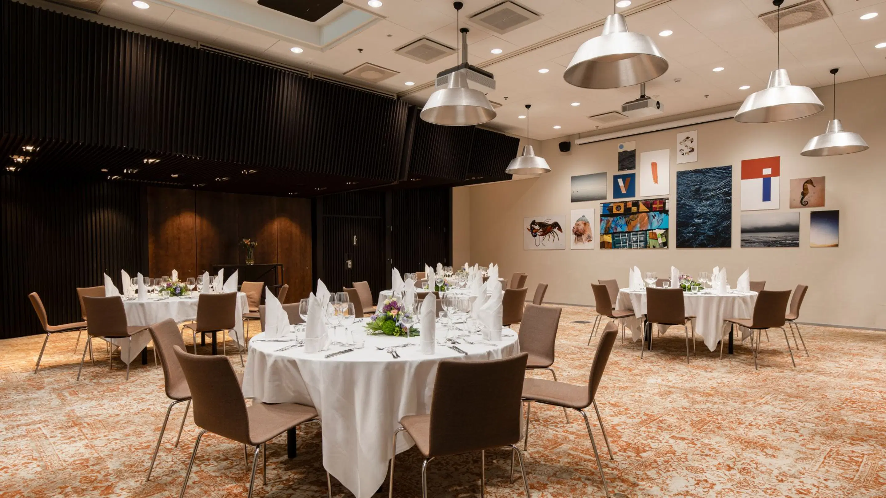 Restaurant dining room with round tables and white tablecloths. Art pieces on wall, high ceiling with pendant lights. Floor has orange-white patterned carpet.