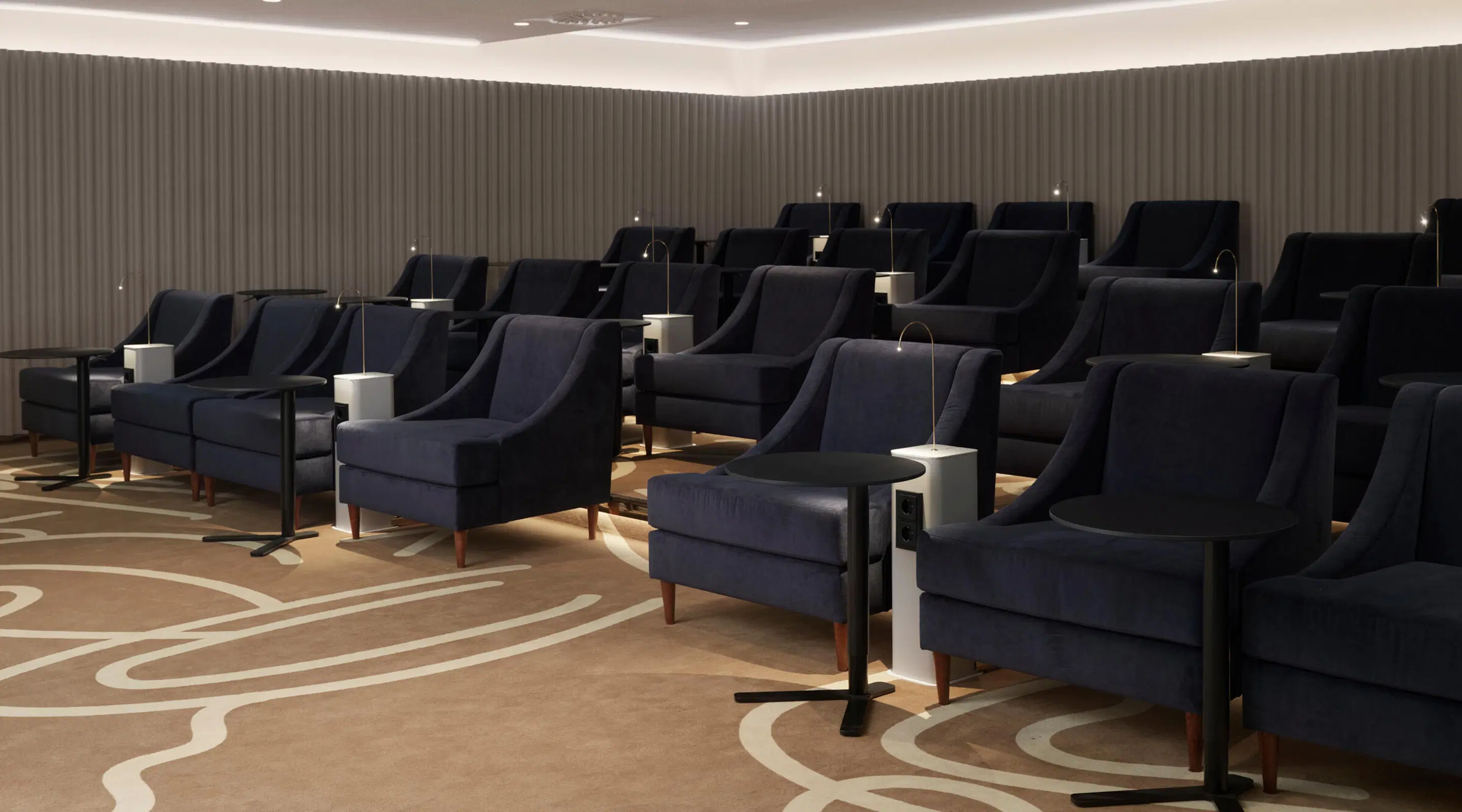 Modern auditorium med mörkblå fåtöljer, små bord och läslampor mellan sätena. Golvet har ett mönstrat mattklätt golv.