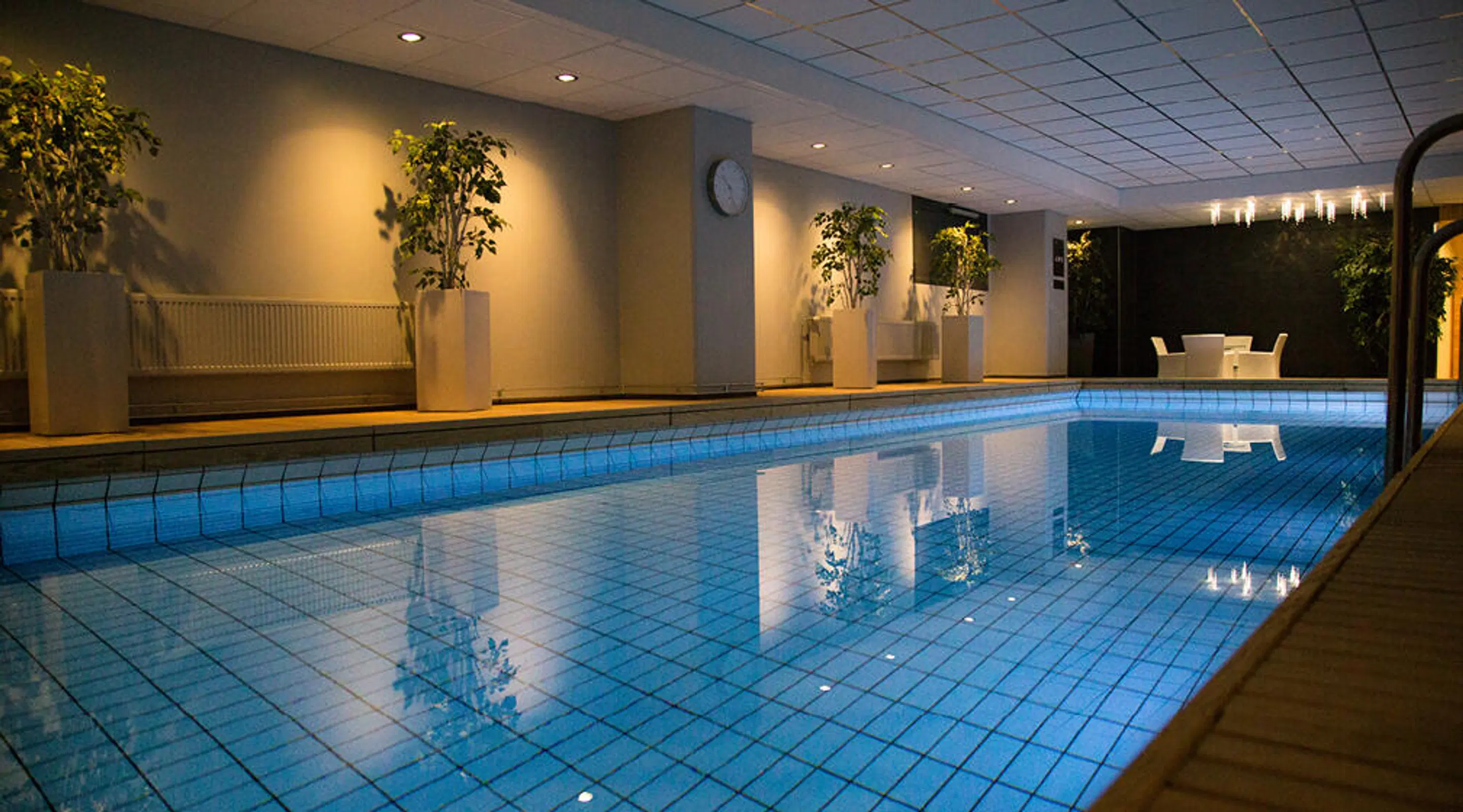 Indoor swimming pool lined with white planters containing greenery. Seating area with table and chairs visible in background. Soft lighting creates calm atmosphere.