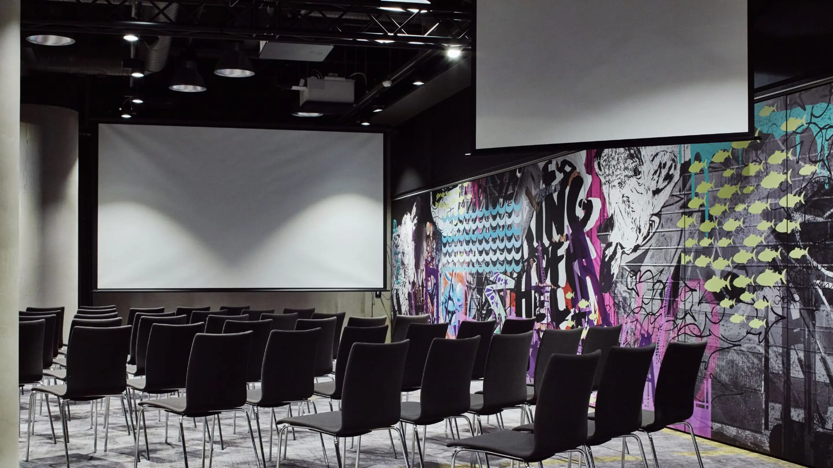 Modernt auditorium med svarta stolar, projektorduk och färgglada graffitiaktiga väggmålningar. Plant golv och god belysning.