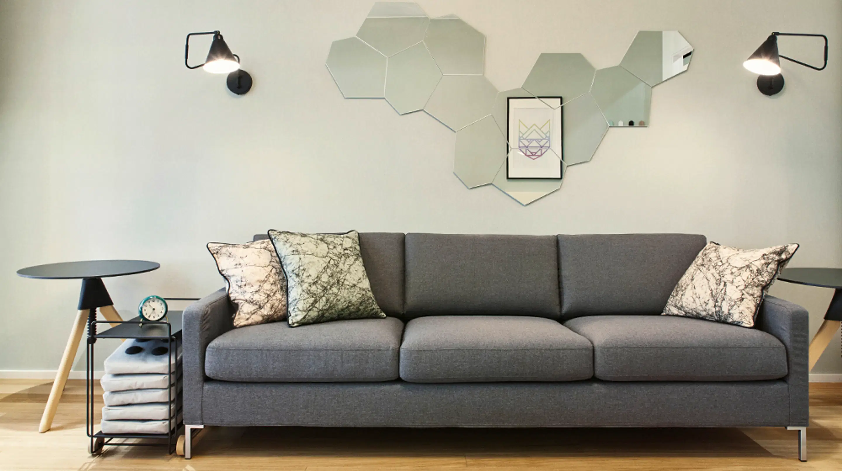 Modern living room with gray sofa, patterned cushions, hexagonal mirrors on wall, and wooden flooring.