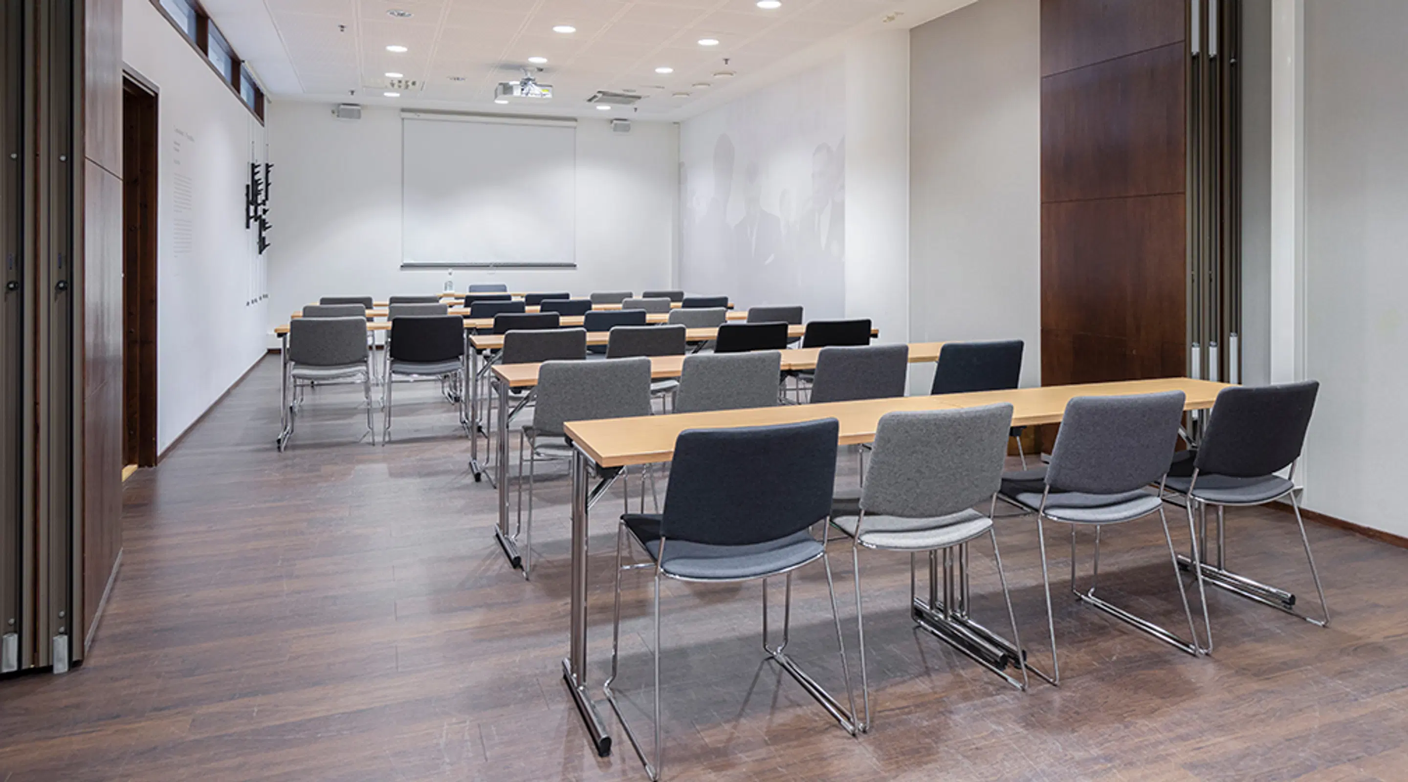 Modern meeting room with tables and chairs arranged in rows, projection screen at front and wooden flooring. Bright space with dark doors on the side.