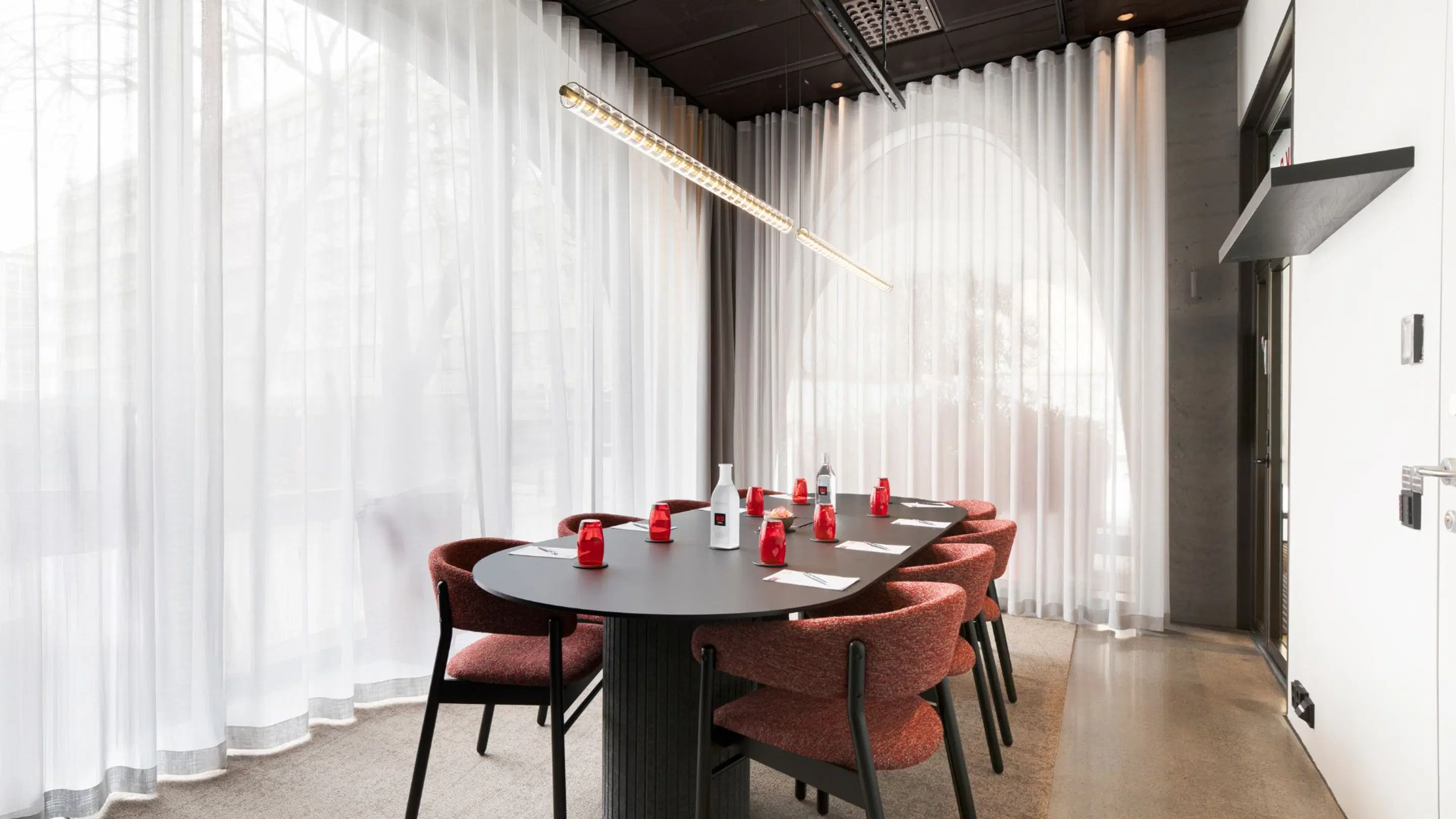 Modern meeting room with oval black table, red chairs, white curtains and beige floor. Red water bottles on the table.