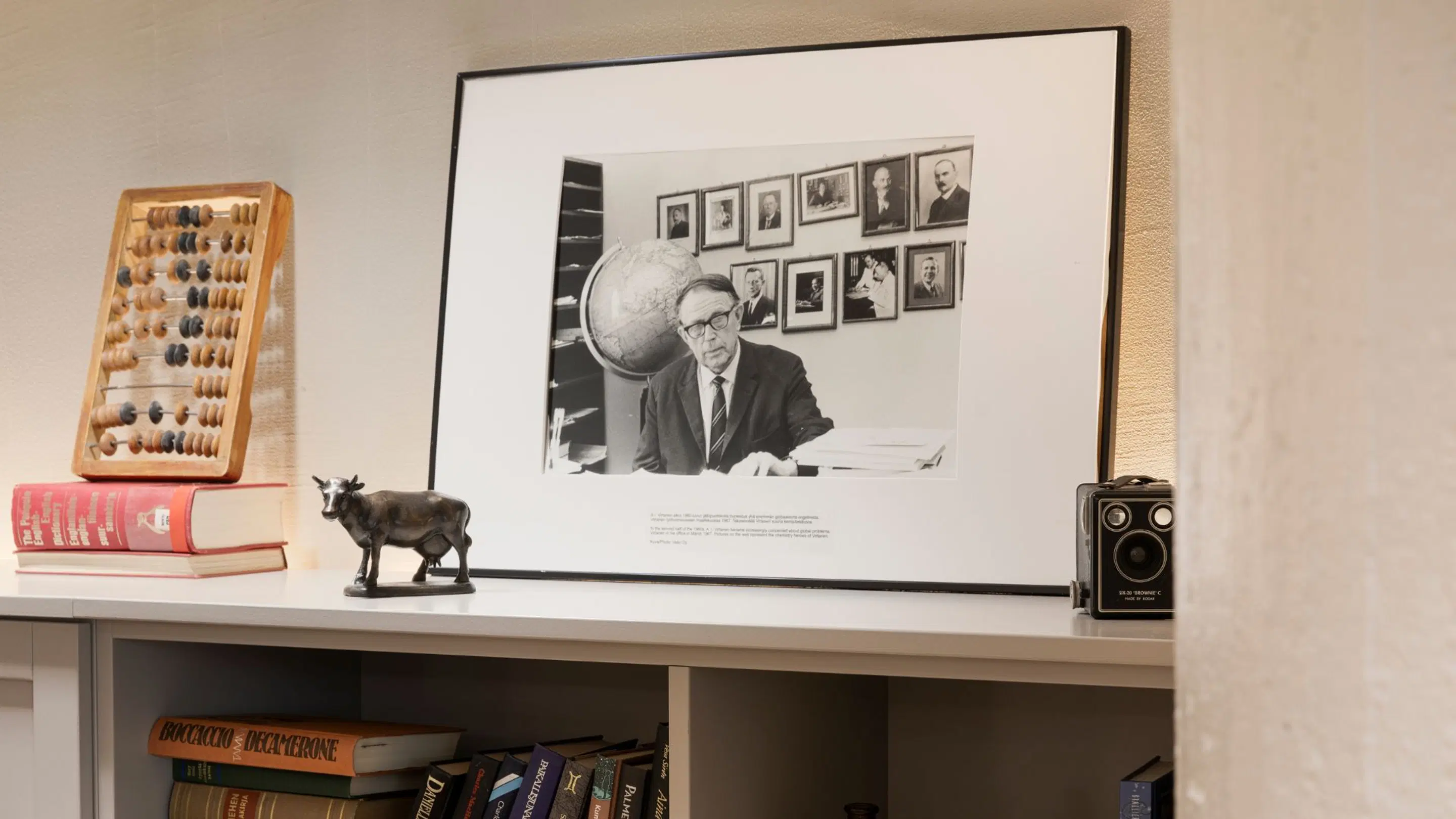 En vit hylla med ett historiskt svartvitt fotografi, en kulram, en liten kostaty och en vintage kamera. Bokhylla under.