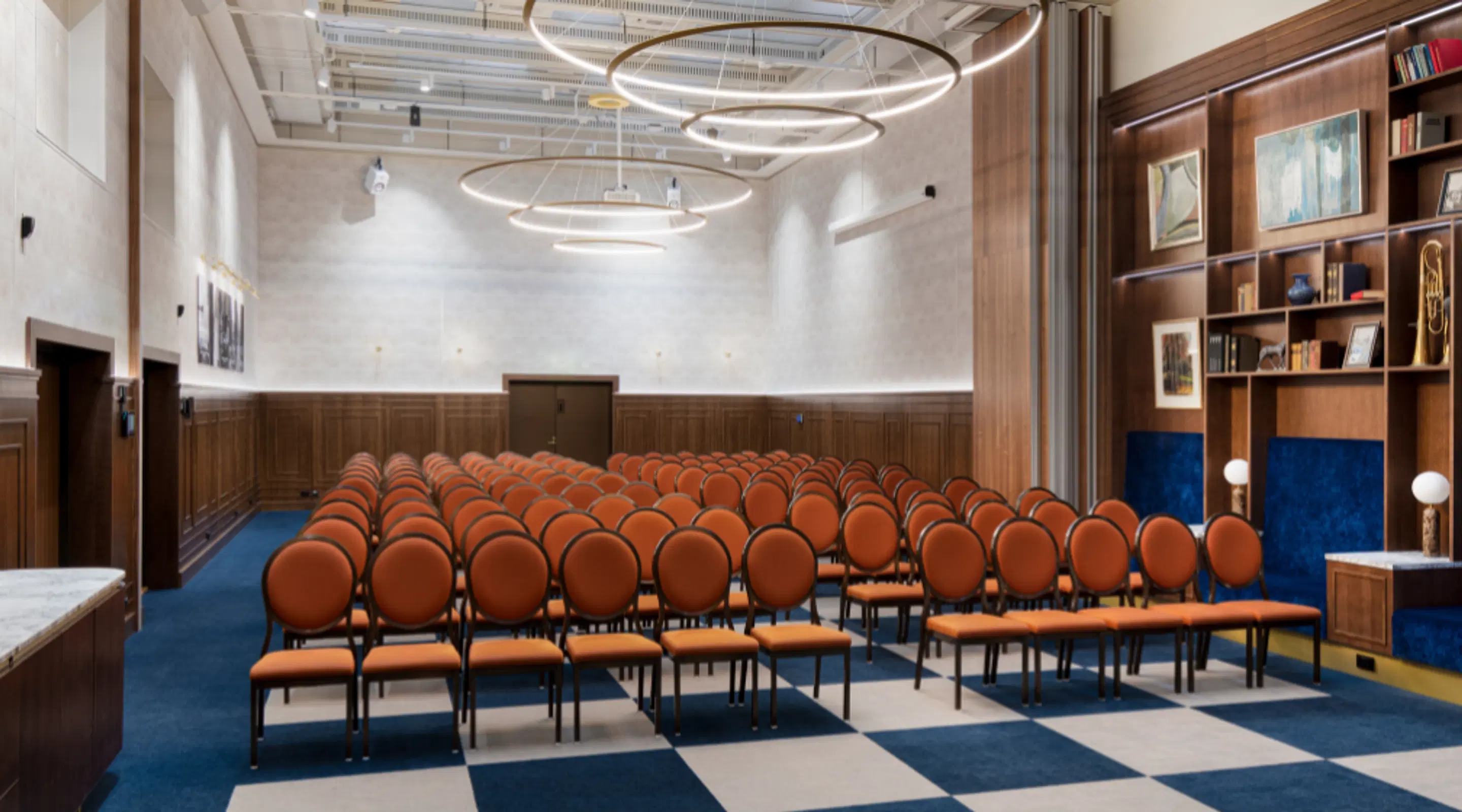 Moderni auditorio, jossa rivistöissä oransseja tuoleja, sininen matto, puiset seinäpaneelit ja kattovalaistus rengasmuodossa.