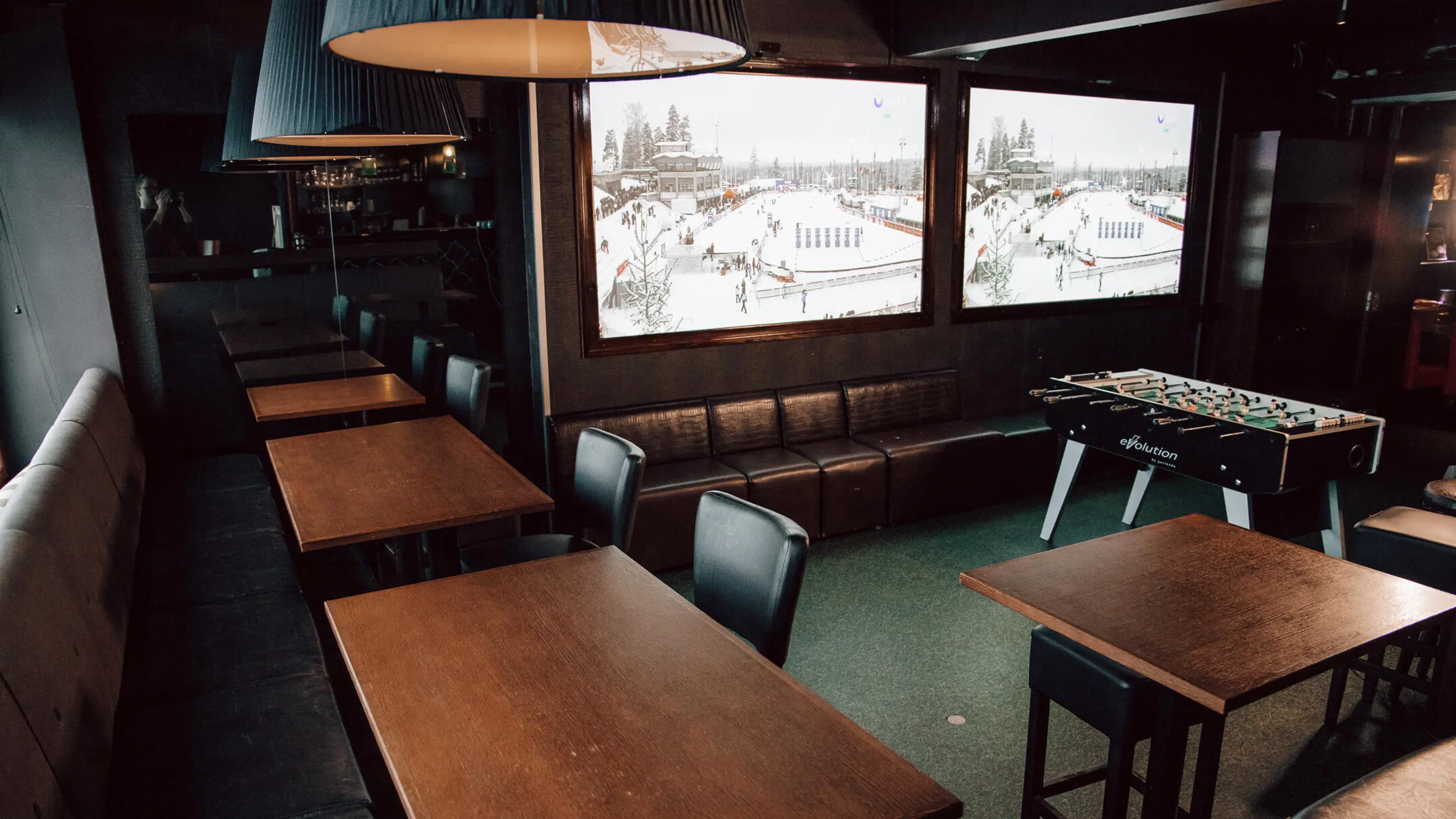 Dimly lit bar with wooden tables, leather chairs and a bench along the wall. Two large screens and a foosball table. The floor has thin green carpet.